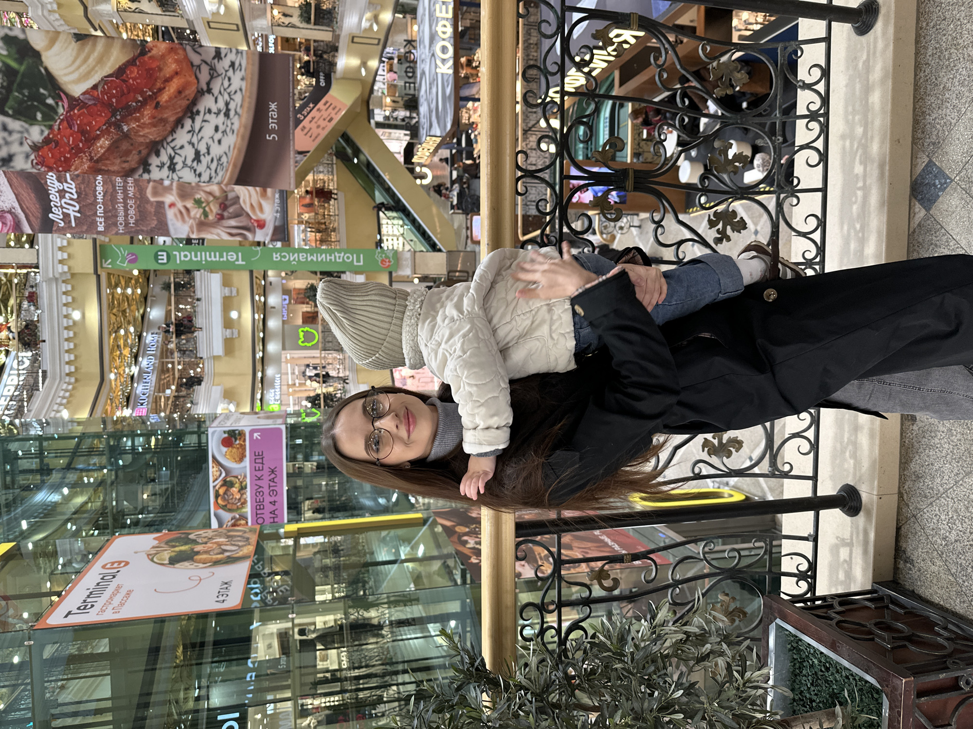 An image, rotated 90 degrees counter-clockwise, depicts a woman holding a baby or toddler within a multi-story shopping mall, likely in Yekaterinburg, Russia. The woman, with long dark hair and glasses, wears a black coat and a grey turtleneck, looking towards the camera with a soft smile. The child, dressed in a light quilted jacket and a beige knitted hat, is held facing away from the camera. They are positioned next to a decorative black metal railing with gold accents, overlooking the lower levels of the mall.
The background reveals a bustling commercial environment with multiple floors, escalators, and various shopfronts. Large advertisements and signs in Russian are prominent. Visible text includes "5 этаж" (5th floor), "4 этаж" (4th floor), and "3 этаж" (3rd floor), indicating different levels. Signage for "Terminal E" and "Terminal гастромаркет в Пассаже" (Terminal gastromarket in Passage) suggests a food court or market area. Other texts like "Поднимайся" (Go up/Ascend), "ОТВЕЗУ К ЕДЕ НА 4 ЭТАЖ" (I'll take you to food on the 4th floor), "Легенды Юга" (Legends of the South), "ВСЁ ПО-НОВОЙ" (All new), "НОВЫЙ ИНТЕР" (New interior), "НОВОЕ МЕНЮ" (New menu), and "КОФЕ" (Coffee) are visible, along with a "KUCHENLAND HOME" sign. The scene appears to be well-lit, suggesting daytime or bright indoor lighting. Green leafy plants in a planter add a touch of nature to the indoor setting.