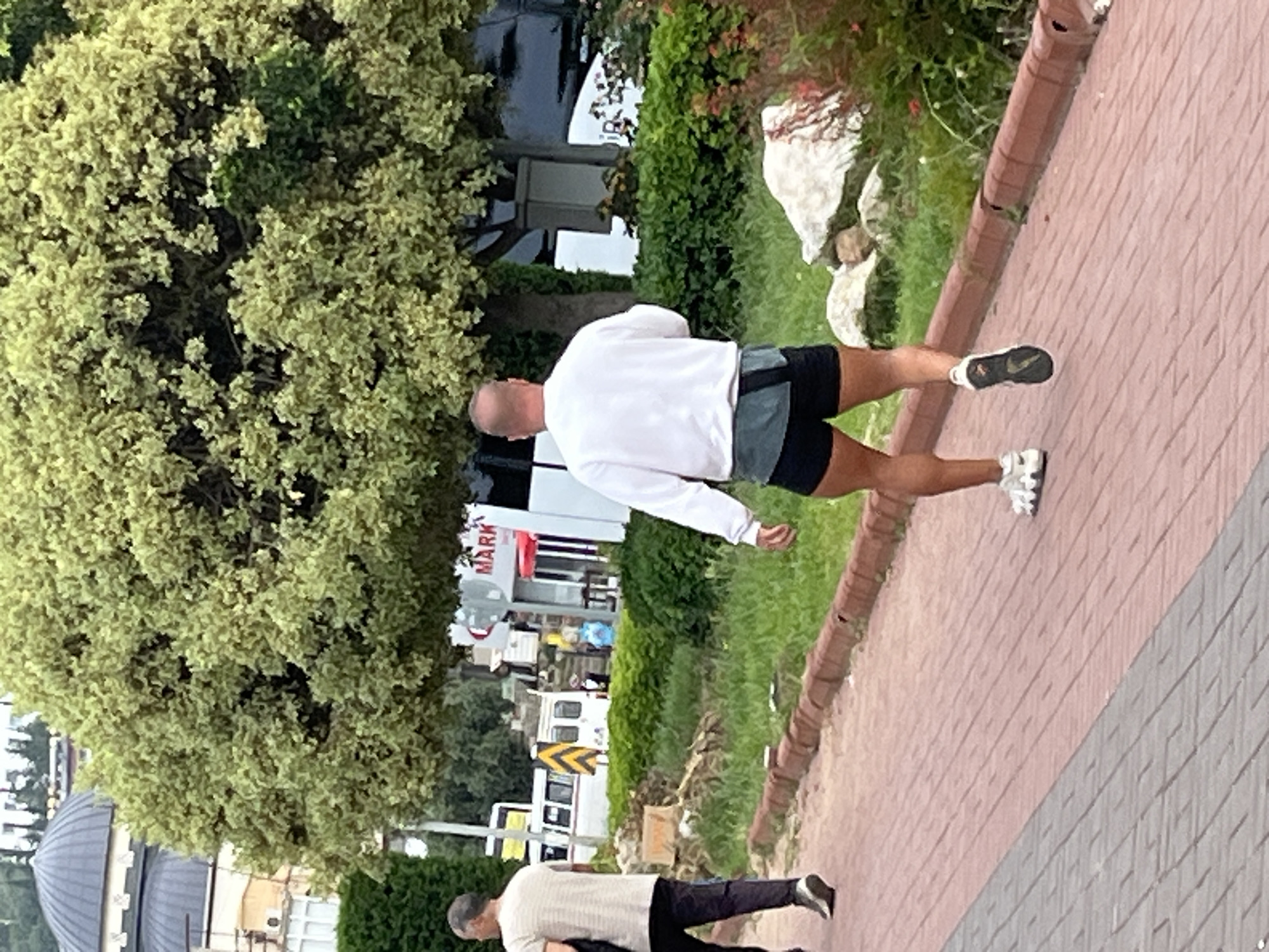 The photograph depicts two men walking on a brick path in Alanya, Turkey.
The first man, facing away from the camera, is wearing a white long-sleeved shirt and black shorts. He is running along the side of the brick path in sneakers with a black and white sole.
The second man, located further down the path, is wearing a light-colored short-sleeve shirt and black pants. The second man is walking in sneakers.
The path is bordered by grass and shrubbery on the left. Behind them, a tree dominates the upper left portion of the frame. In the background, there is a building with an arched roof, along with street signage. A building with a "MARK" sign is also visible.