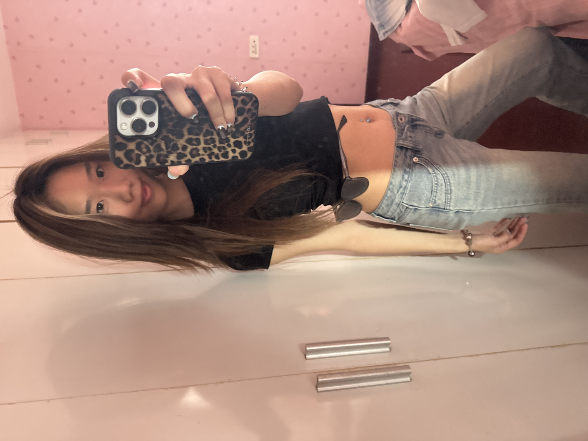 A young woman is taking a selfie in a room with pink walls decorated with small white hearts. She is wearing a black crop top, light blue jeans, and has a belly button piercing. She is holding a phone with a leopard-print case in her left hand. Sunglasses are tucked into the waistband of her jeans. There are dresser drawers in the bottom of the frame. Based on the setting and the presence of a standard wall outlet, the photo may have been taken in a private residence. There is no information available in the image to confirm that this photo was taken in the Shapingba District of China.