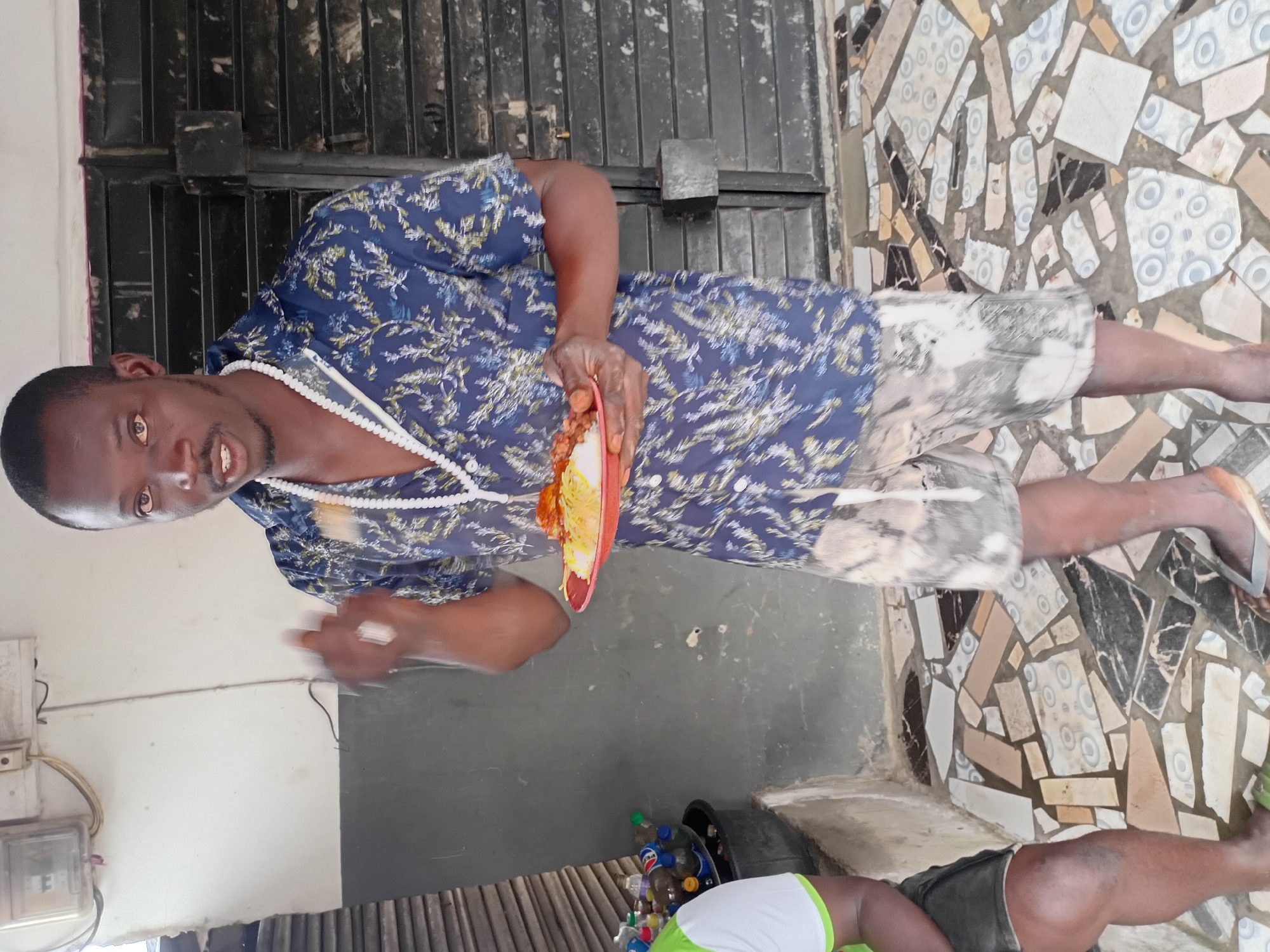 A dark-skinned man stands holding a plate of food and gestures with his left hand. He wears a blue patterned short-sleeved shirt and patterned shorts. A white beaded necklace hangs around his neck. He is standing on a floor with a mosaic tile pattern. In the background, to the left, is a person sitting down, wearing a green and white shirt and dark shorts. Behind them is a collection of plastic bottles. To the right of the man is a black, slatted structure, possibly a door or gate. The wall behind the man is light-colored, with some dark marks and a white electrical box mounted on it. The overall setting appears to be an outdoor or semi-outdoor area, possibly a patio or porch.