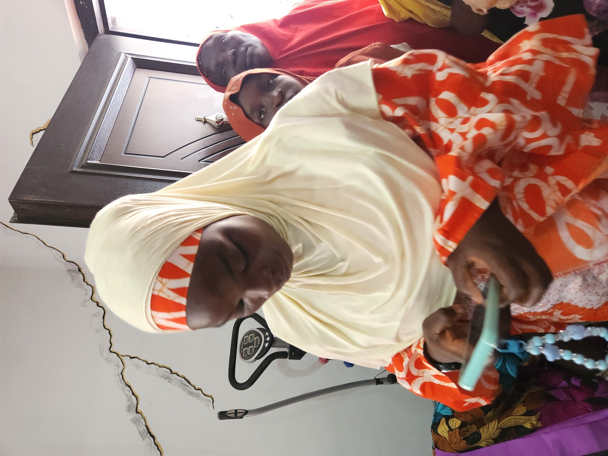 The image depicts a scene within a home in Sabon Tasha, Nigeria. In the foreground, a woman wearing a light yellow hijab with an orange and white patterned band is looking down at a teal smartphone she is holding. She is also wearing an orange and white patterned garment. To her right, a young girl in an orange hijab is partially visible, and behind her, an older woman in a red hijab is looking towards the camera. In the background, a dark brown door is visible, and to the left, a white wall with a black and grey abstract design is present. A piece of exercise equipment, possibly a stepper, is also in the background to the left. The overall impression is of an indoor gathering or moment of personal interaction within a residential setting.
