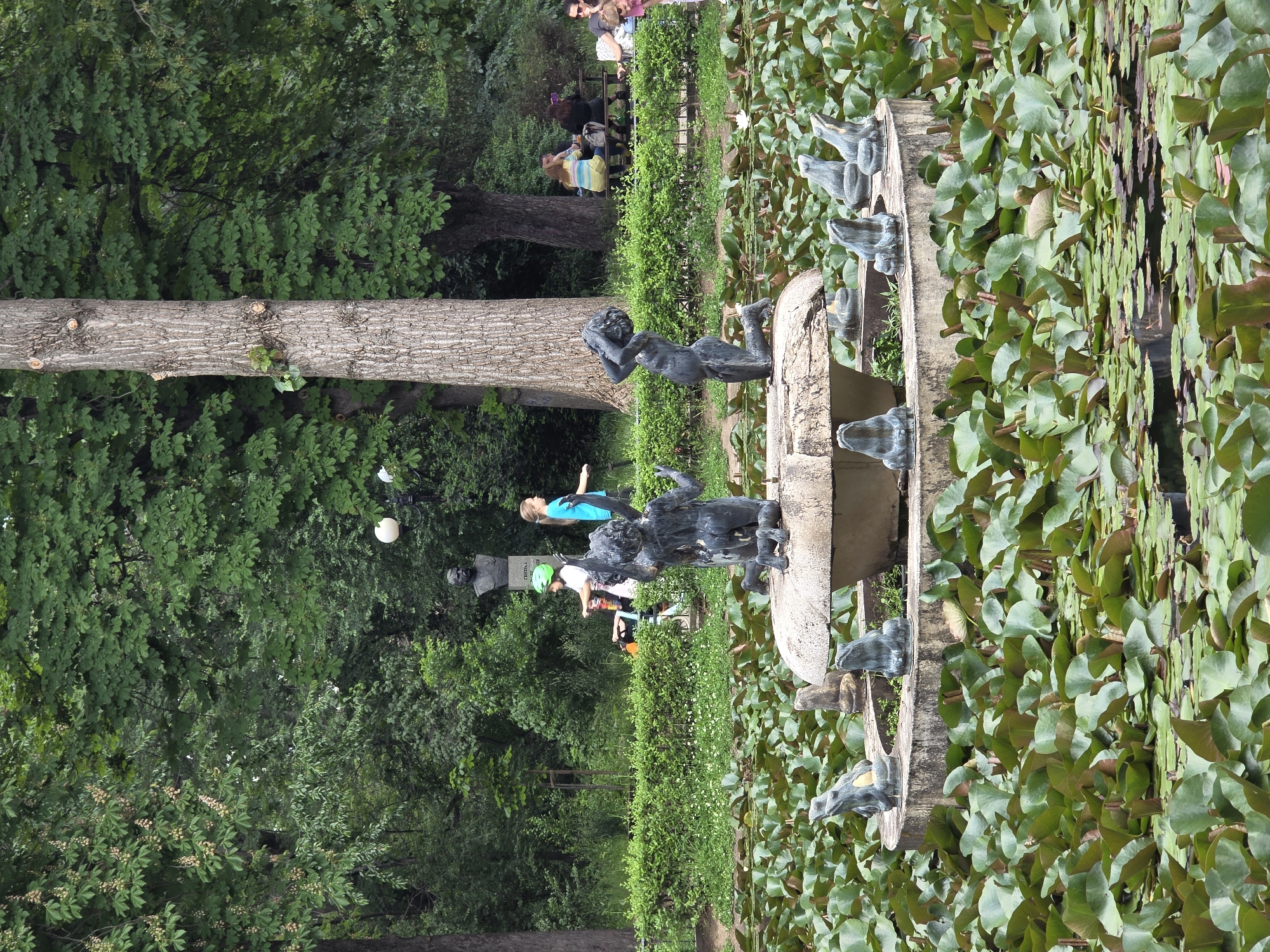 The image depicts a water fountain in a park in Sofia, Bulgaria. The fountain is circular with several bronze figures, including children and frog-like creatures. The fountain is surrounded by a pond filled with lily pads. Several people, including a woman in a blue shirt, a child with a green helmet, and others on a bench, are in the background. Trees and greenery are visible throughout the scene. A tree trunk is noticeable on the left side of the frame.