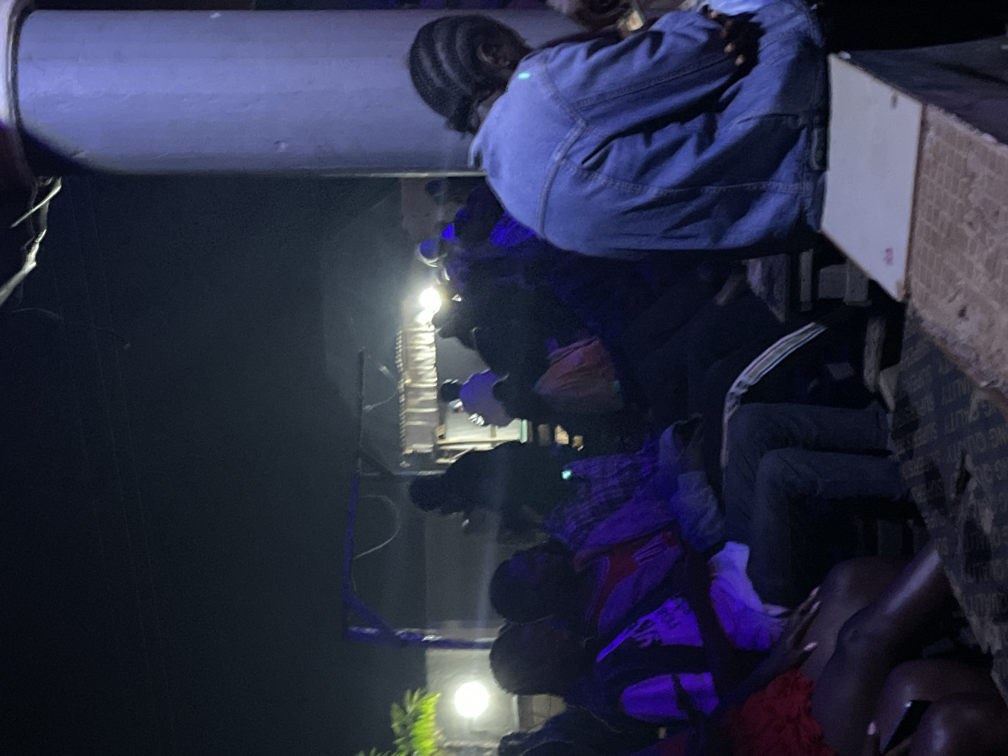 In the city of Durumi, Nigeria, a group of individuals are gathered outdoors at night. The scene is dimly lit by artificial lights, casting a purplish hue on some of the subjects. In the foreground, a person wearing a denim jacket is seated, with their back to the camera, facing away from the viewer and towards the rest of the gathering. To the left, several other people are seated, some with their arms around each other, suggesting a communal or social setting. In the background, more figures are visible, some appearing to be standing or moving, with a structure that looks like a makeshift stage or platform. A bright light illuminates a portion of the background, possibly from a stage light or another source of illumination. The ground appears to be uneven and covered with some materials, possibly for seating or as a foundation. The overall atmosphere suggests an event or gathering taking place in an informal, outdoor setting.