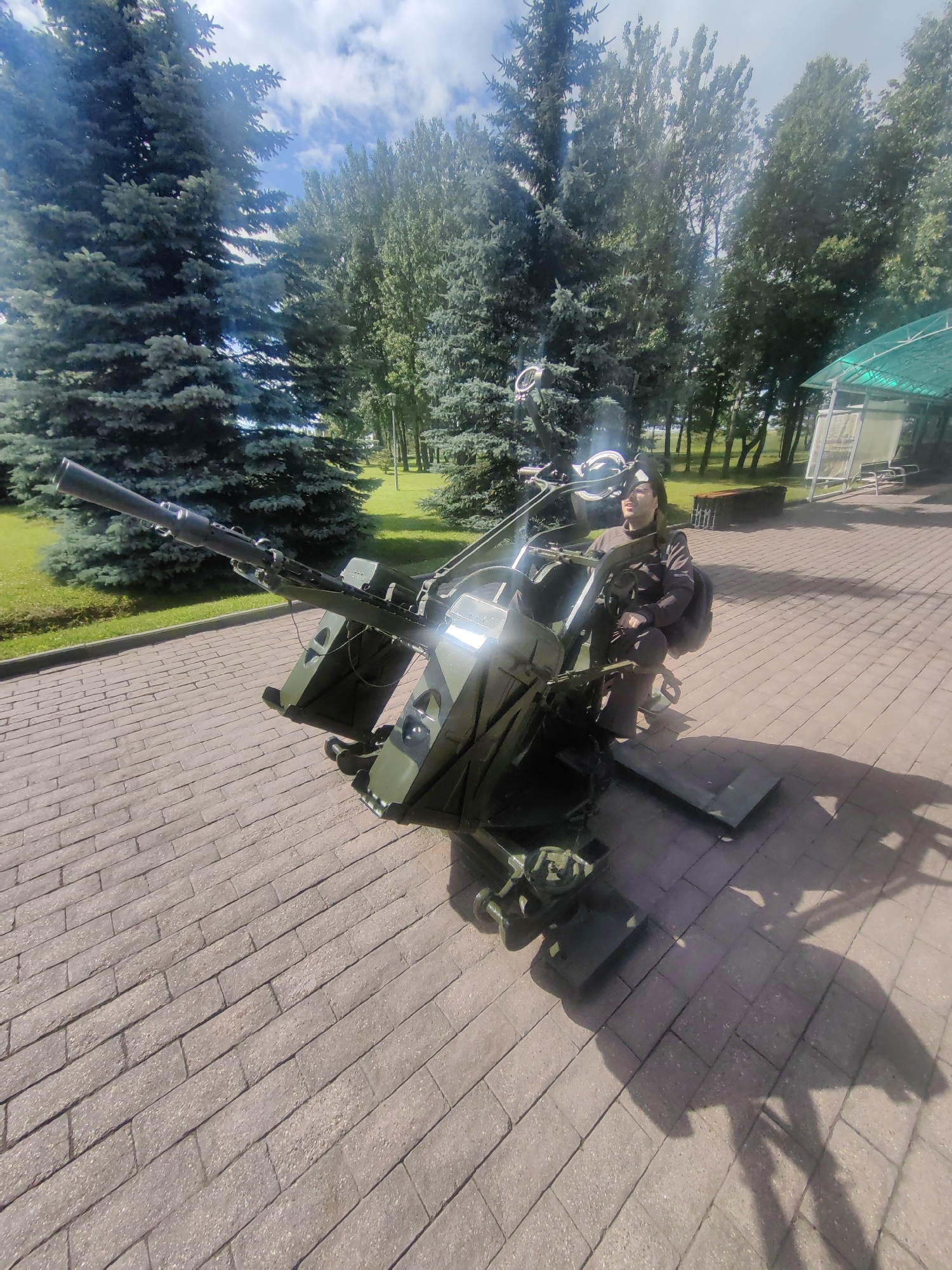 In Kubinka, Russia, a person is seated on a vintage anti-aircraft gun display, which is positioned on a brick-paved area. The gun is dark green and appears to be in a static exhibit. The person, wearing glasses and a dark jacket, is looking to the right. Behind them, lush green trees and a bright, cloudy sky form the backdrop. A green awning covers a sheltered area with seating in the background. The scene suggests an outdoor museum or park setting, possibly related to military history.