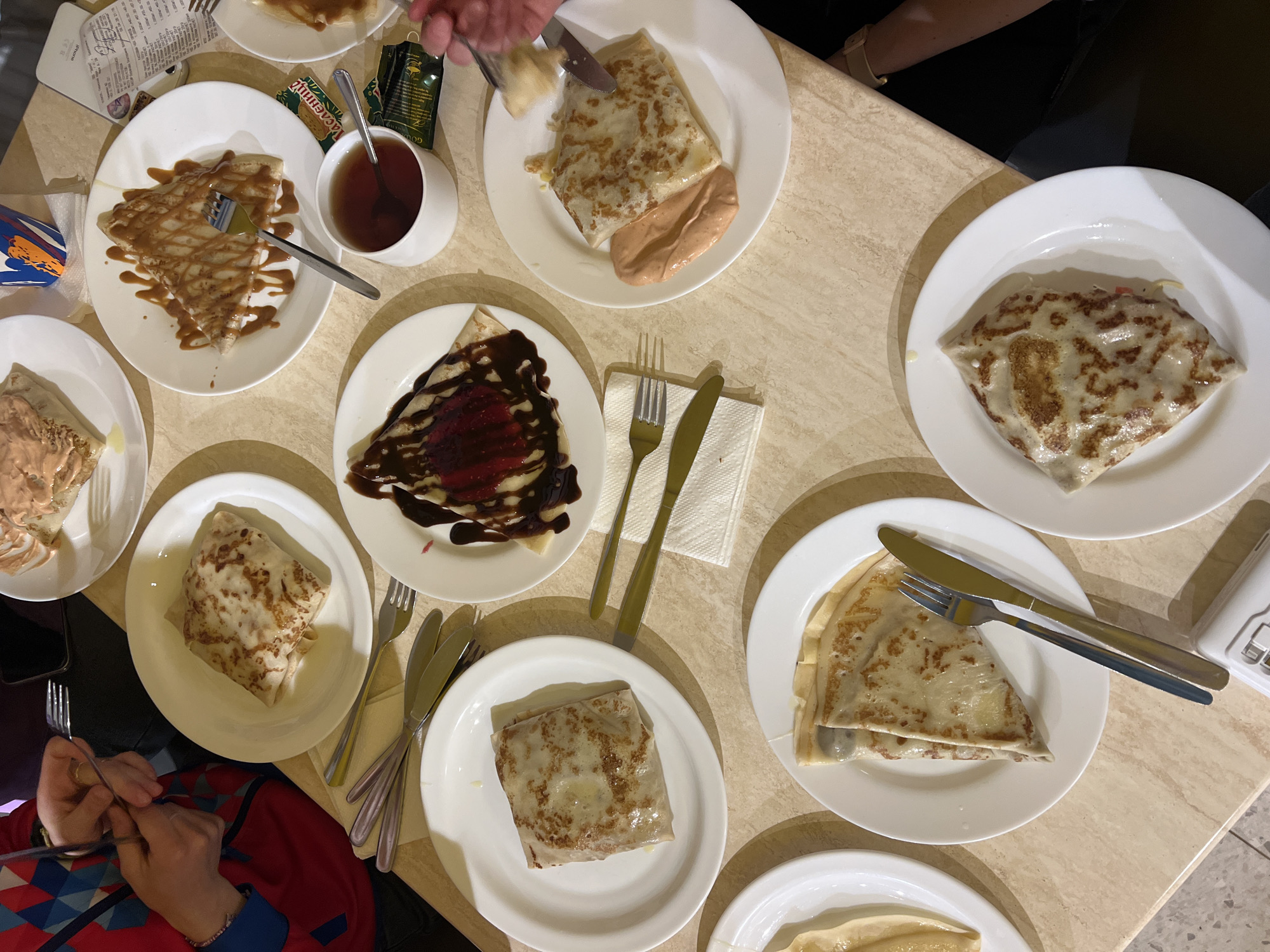 The image shows a table setting with multiple plates of crepes and other items. A cup of tea sits next to a plate of crepes with caramel sauce. Other plates show crepes with what appears to be meat filling, chocolate sauce and strawberry filling. Several sets of utensils are positioned around the plates. There are at least three visible people, two with hands and/or arms visible and one with both hands visible. One person is holding a fork and knife. The location is likely a restaurant or dining establishment, but not identifiable to Berezniki.