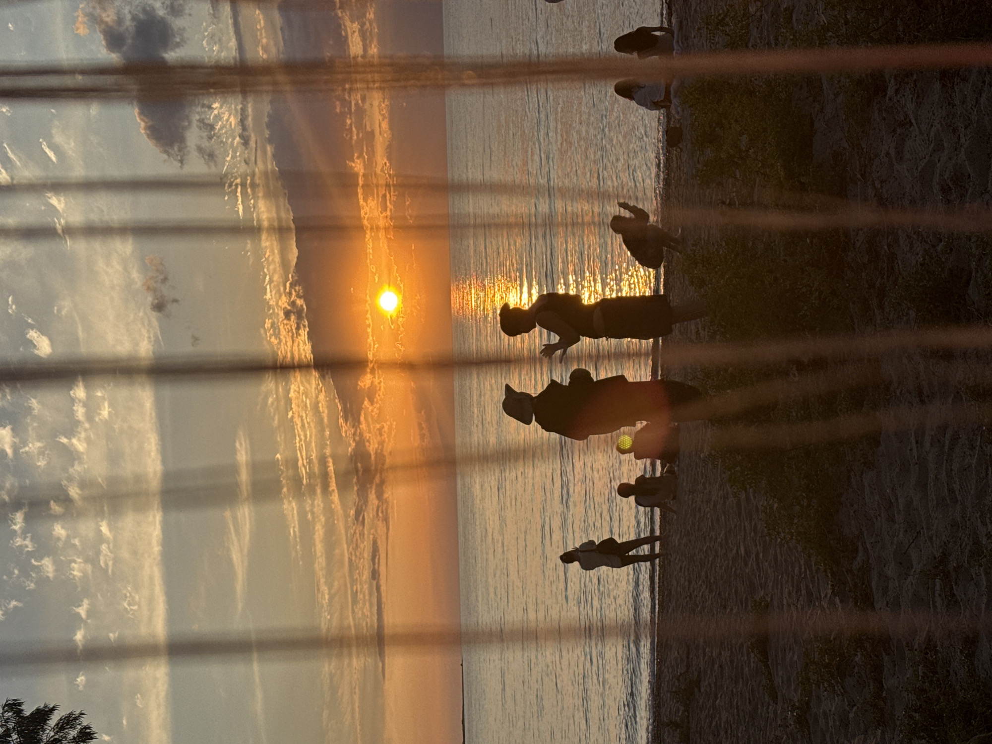 A vertical, full shot captures a sunset over a body of water at Basbeshik, Kyrgyzstan. The sun, a bright orange disc, is positioned slightly right of the center of the horizon. Its reflection creates a shimmering path across the calm, blue water. The sky above is a gradient of warm oranges and yellows, transitioning into blues and whites with scattered, wispy clouds. In the foreground, a sandy beach is visible with several people in silhouette. Two individuals stand near the water's edge, facing each other, their figures darkened against the bright sky. Further back, other people are seated or crouching on the sand. The scene is partially obscured by vertically oriented, translucent strands, possibly a curtain or netting, creating a layered effect with vertical shadows. The overall atmosphere is tranquil and serene, characteristic of a late summer evening.