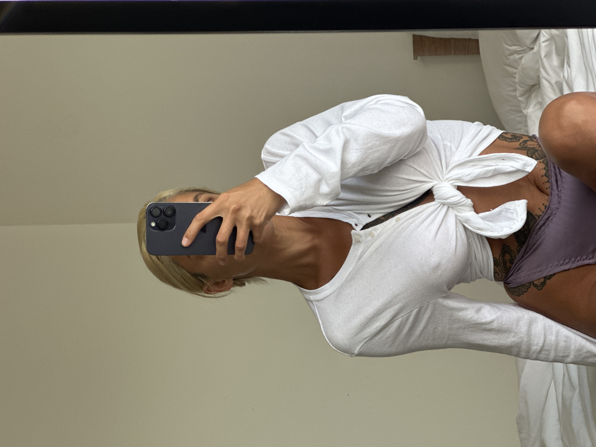 A person with blonde hair takes a mirror selfie while sitting on a bed. They are wearing a white long-sleeved shirt, tied at the waist, and purple underwear. The person is holding a black smartphone to obscure their face. The background is a plain wall and a bed with white linens. A wooden headboard is visible on the right side of the image, and a mirror reflects the scene. The location is Kuta Selatan, Indonesia.