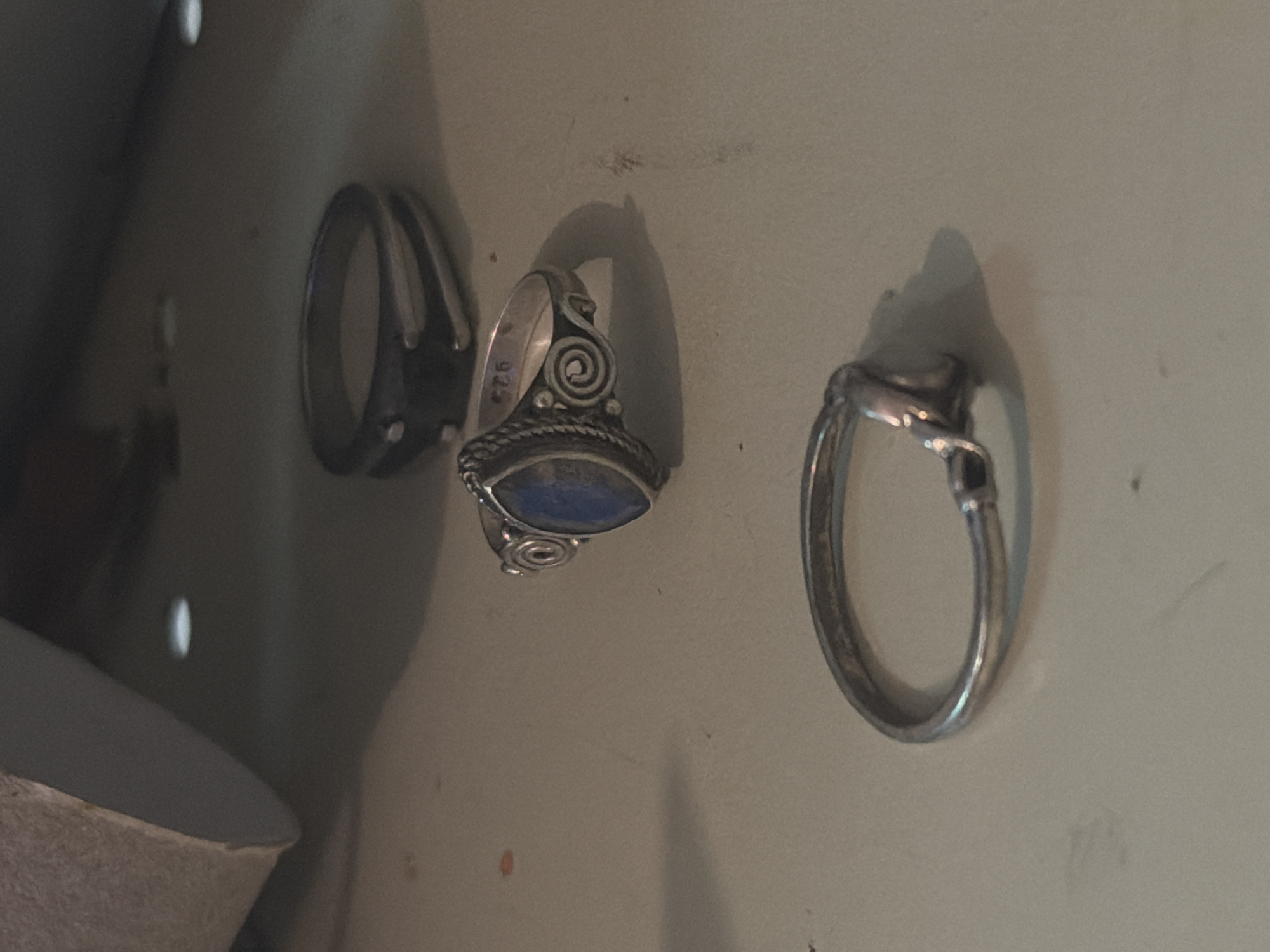 Three silver rings are arranged on a light-colored surface. The ring in the foreground is simple with a twisted band. The middle ring features a marquise-shaped blue stone and decorative spirals on the band, with the inscription "925" visible. The ring in the background is darker and has a geometric design. The surface appears to be indoors. There is no discernible information indicating a location in Tel-Aviv, Israel, or any specific action or interaction.