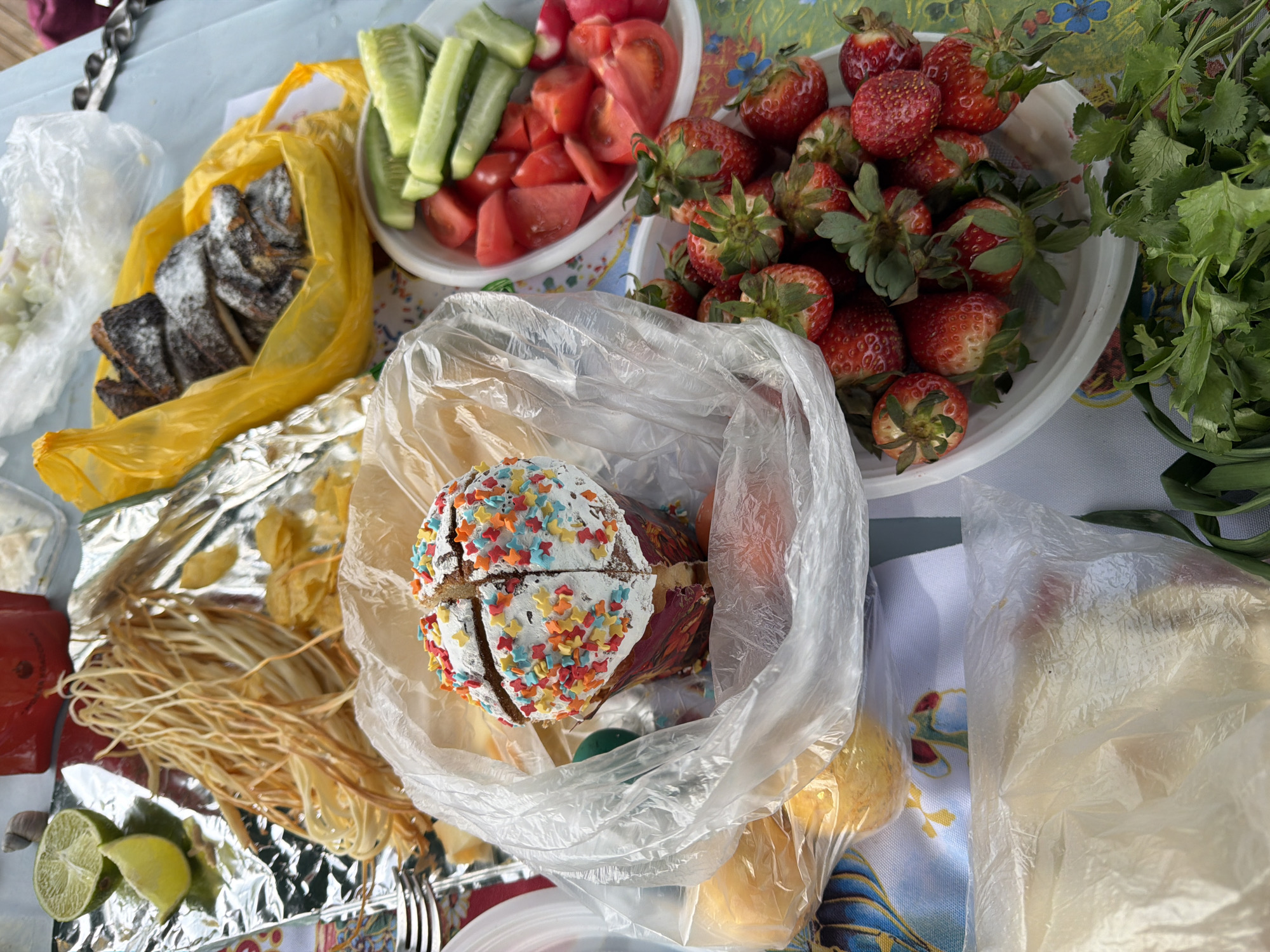 The image shows a table laden with food, likely taken in Olginskaya, Russia. There is no indication of people, actions, or interactions in this image. The objects are diverse, including:
* A cake decorated with colorful sprinkles, partially sliced and in a clear plastic bag.
* Strawberries in a white bowl with green tops.
* Sliced cucumbers and tomatoes in a white bowl.
* String cheese on foil.
* A bag of what appears to be pastry.
* Lime wedges.
* Fresh cilantro.
* Several other obscured items in plastic bags.
* Cutlery
The environment is a tabletop setting, with a mix of white tablecloth and bare table visible.