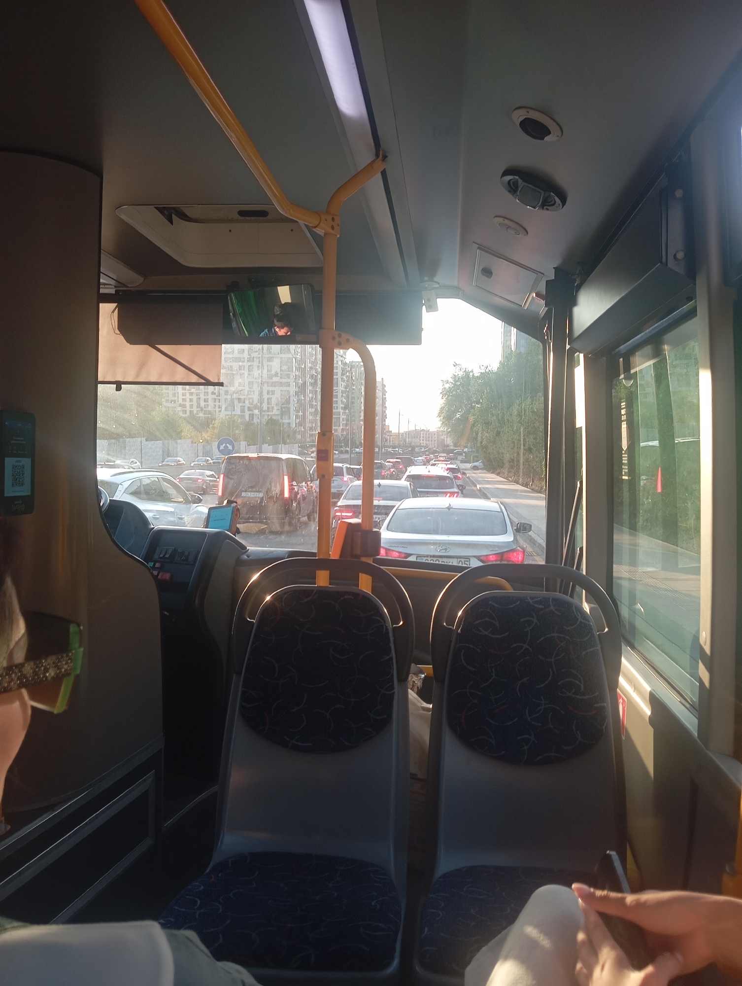 The image is taken from inside a bus looking forward, showing a traffic jam on a city street. The bus has several rows of seats visible, with patterned upholstery. The dashboard and driver's area are partially visible in the foreground. Outside the bus, a long line of cars is stopped on the road, with buildings and trees lining the street. The lighting suggests it is daytime, with sunlight filtering through the windows. The license plate of a silver Hyundai sedan visible on the right side of the road appears to be in Cyrillic script, consistent with Kazakhstan. The presence of similar license plates on other vehicles further supports this location. The overall scene depicts urban traffic congestion in Almaty, Kazakhstan.