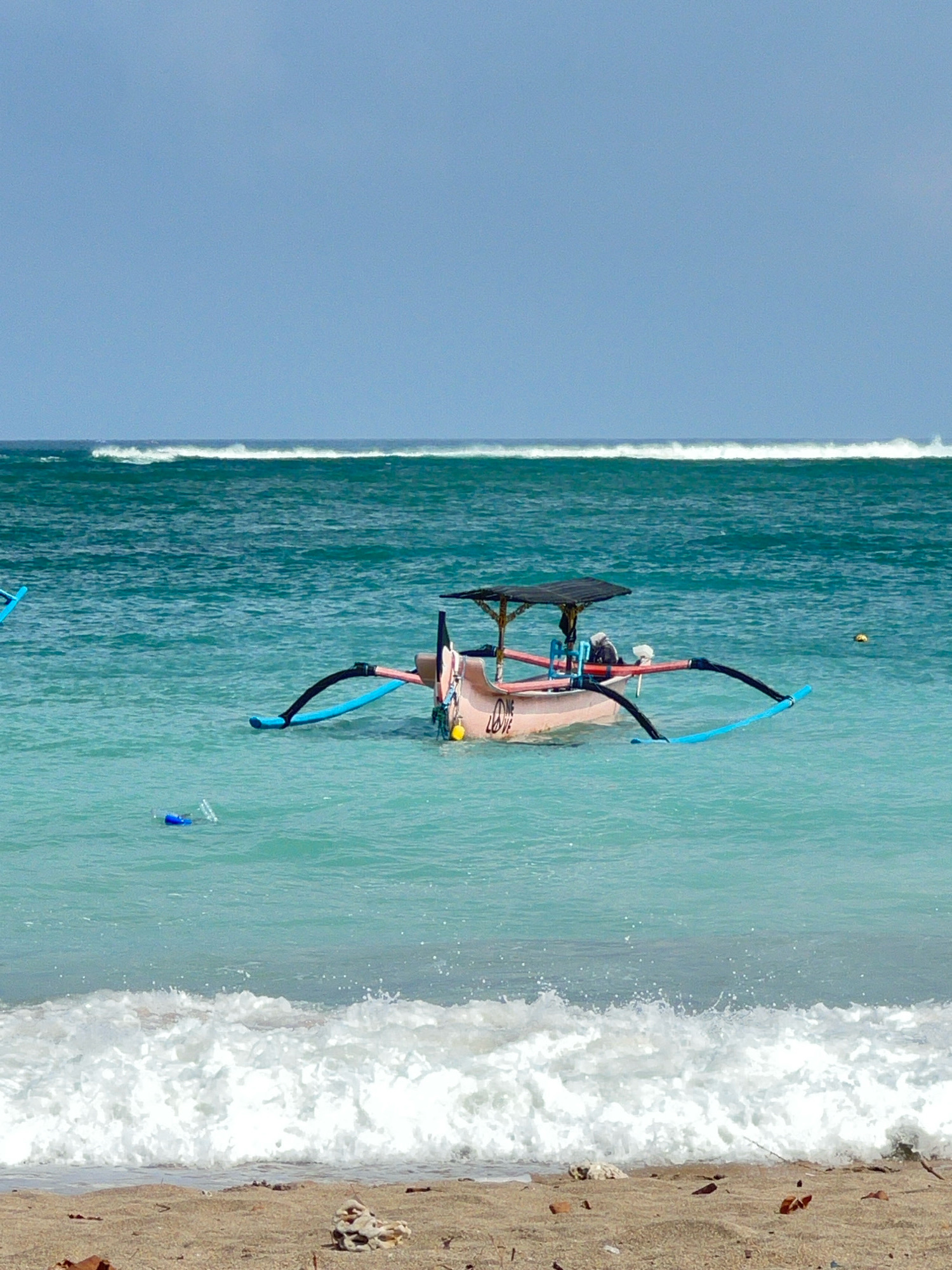 An offshore, narrow, outrigger boat with a black canopy floats on calm, teal ocean water under a clear, light blue sky. The boat is light pink and has writing on its side. Two long, blue outriggers extend from the sides of the boat, connected by black poles. A single, yellow buoy is visible in the water to the right of the boat. In the middle ground, a gentle wave breaks with white foam. In the foreground, the sandy shore is visible with scattered debris and shells. A blue plastic bottle and its cap are floating in the water to the left of the boat. The scene appears to be a tranquil beach in Kuta, Indonesia.