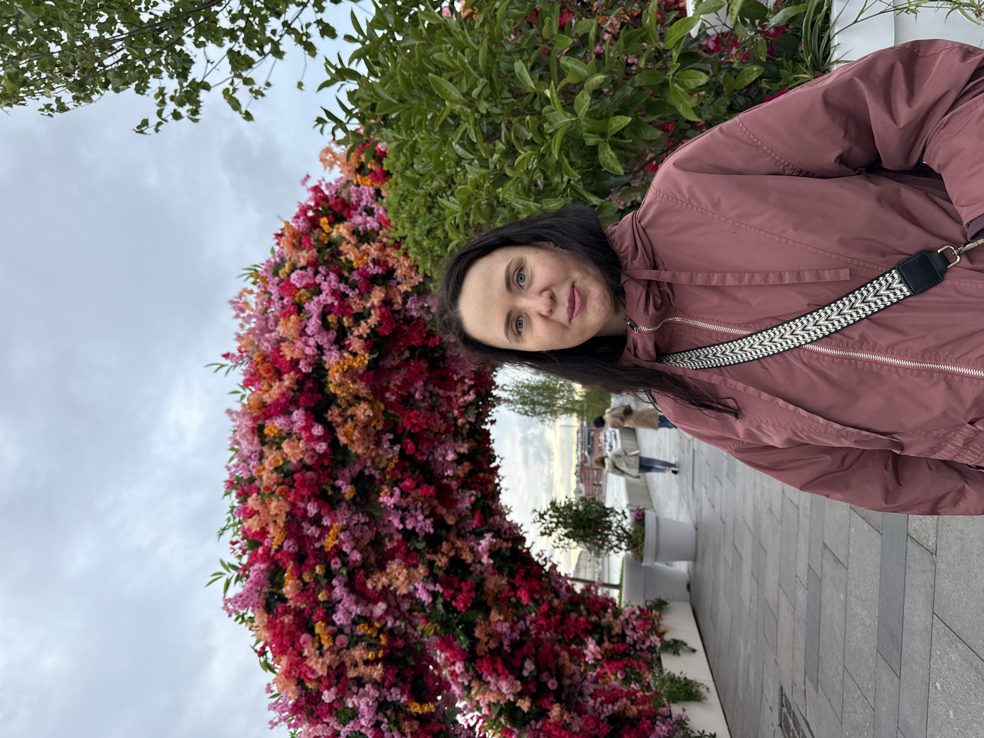 A middle-aged woman wearing a dark mauve hooded jacket and a black-and-white patterned strap stands beside a large floral archway in Zaryadye Park in Moscow, Russia. The archway is covered in a dense arrangement of pink, red, and orange flowers. Behind the archway, other people can be seen walking on a paved pathway. The sky is overcast. The woman is facing the camera with a slight smile.