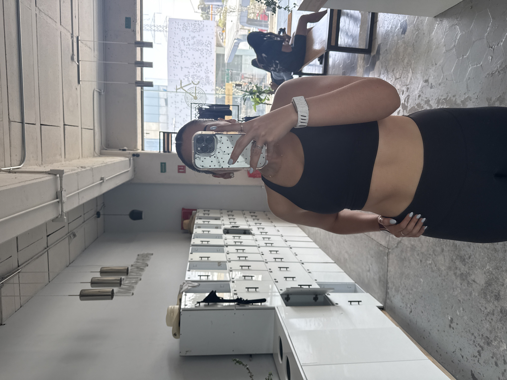 A woman in black athletic attire, including a sports bra and leggings, takes a selfie in a locker room setting. She holds an iPhone with a dotted case. The locker room features rows of white lockers with black handles. Overhead are cylindrical light fixtures suspended from the ceiling. A view outside reveals a city scene with buildings and pedestrians, identifiable as Puebla, Mexico. A woman is sitting near the window in a chair. The floor is made of gray stone.