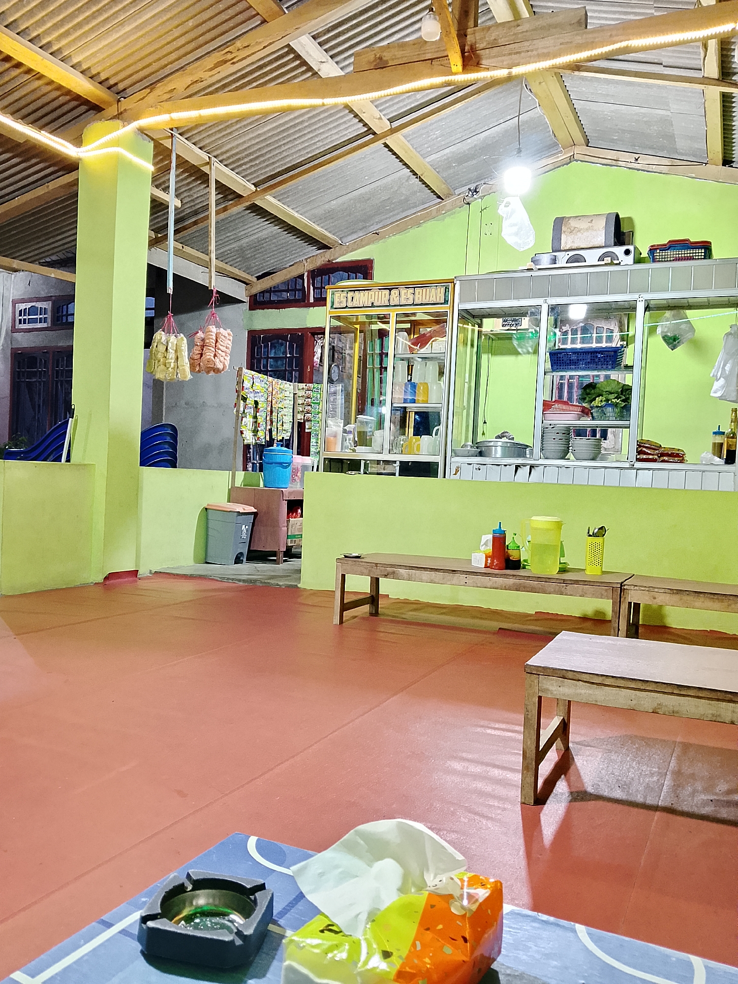 The image depicts the interior of a casual eatery, likely a warung or small restaurant, in Purba Sakti, Indonesia. The space has a vibrant lime green wall and a red floor. The ceiling is constructed with corrugated metal and supported by wooden beams, some of which are adorned with yellow LED strip lights.
In the background, a counter area is visible with shelves displaying various items. These include stacks of plates, bowls, a yellow pitcher, a yellow utensil holder filled with cutlery, bottles of condiments (ketchup and soy sauce), and what appear to be packaged snacks. A gas stove is also present on a shelf. Hanging from above are two bunches of snacks, possibly crackers or fried dough. Adjacent to this area is a display case with more items.
The seating area consists of simple wooden benches. On one of the tables, an ashtray with a small amount of ash and some green substance is visible, along with a partially opened packet of tissues. The overall atmosphere suggests a functional, unpretentious dining establishment, potentially open-air given the roof construction. There are no people visible in the scene.