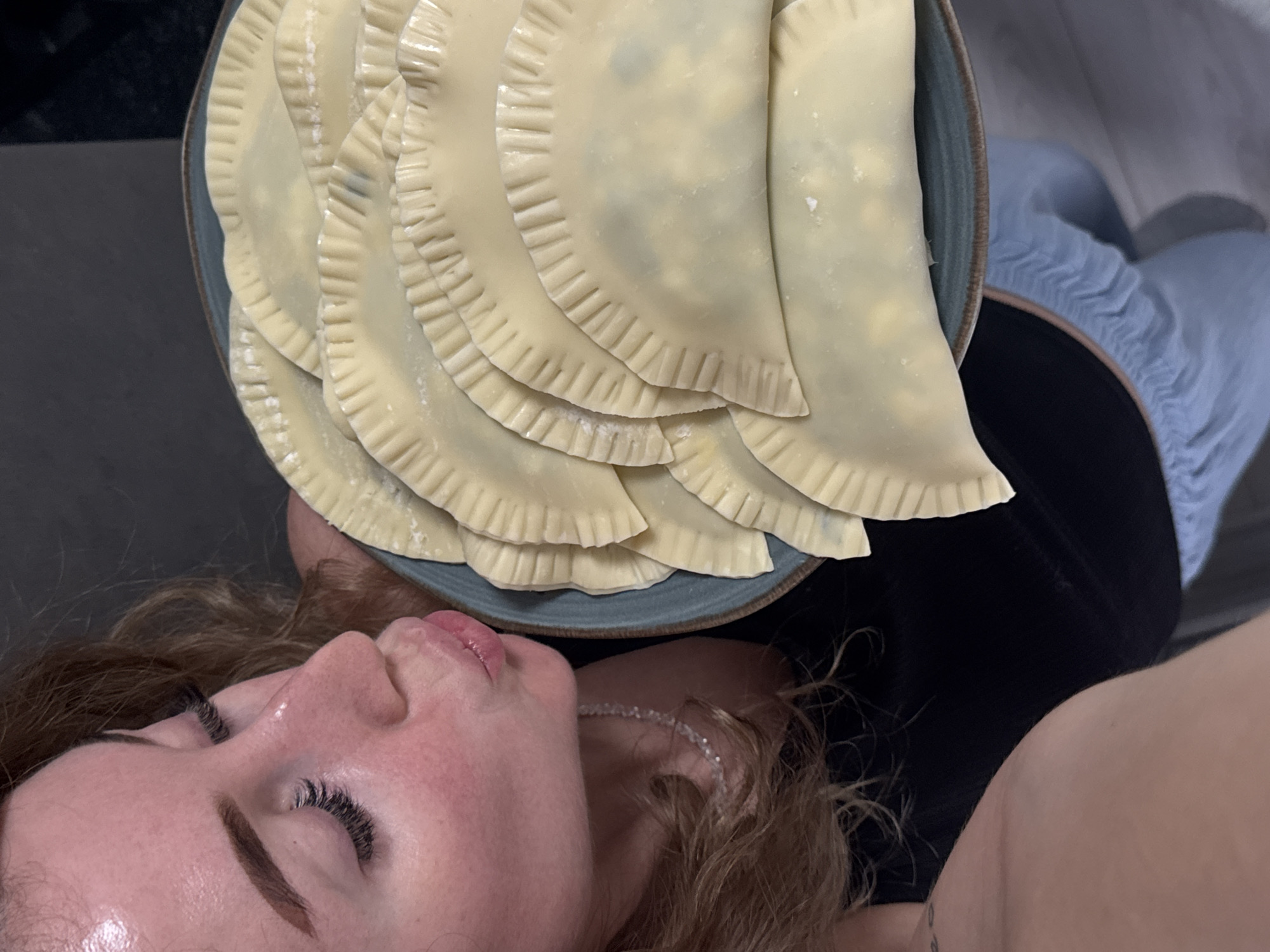 This media file captures a close-up, overhead view of a person holding a plate of uncooked chebureki, a traditional Caucasian fried turnover, likely in a home setting. The plate, a muted blue with a brown rim, is filled with several chebureki that appear to be freshly made, their crimped edges distinct. The dough is a pale yellow, suggesting it is uncooked.
The person holding the plate is positioned in the foreground, with their face turned upwards and eyes closed. Their skin appears flushed, and their lips are slightly parted. They have long, dark eyelashes and their eyebrows are well-defined. A delicate, shimmering necklace is visible around their neck. Their hair is a wavy, light brown.
The background is partially visible, showing a dark surface to the left and a person wearing light blue pants and a black top to the right. The overall lighting suggests an indoor setting, possibly during the day, given the natural light illuminating the person's face. The mood conveyed is one of repose or anticipation, possibly related to preparing or enjoying a meal. There is no visible text in the image. The location context of Chaikovsky, Russia, provides cultural insight into the food being prepared.