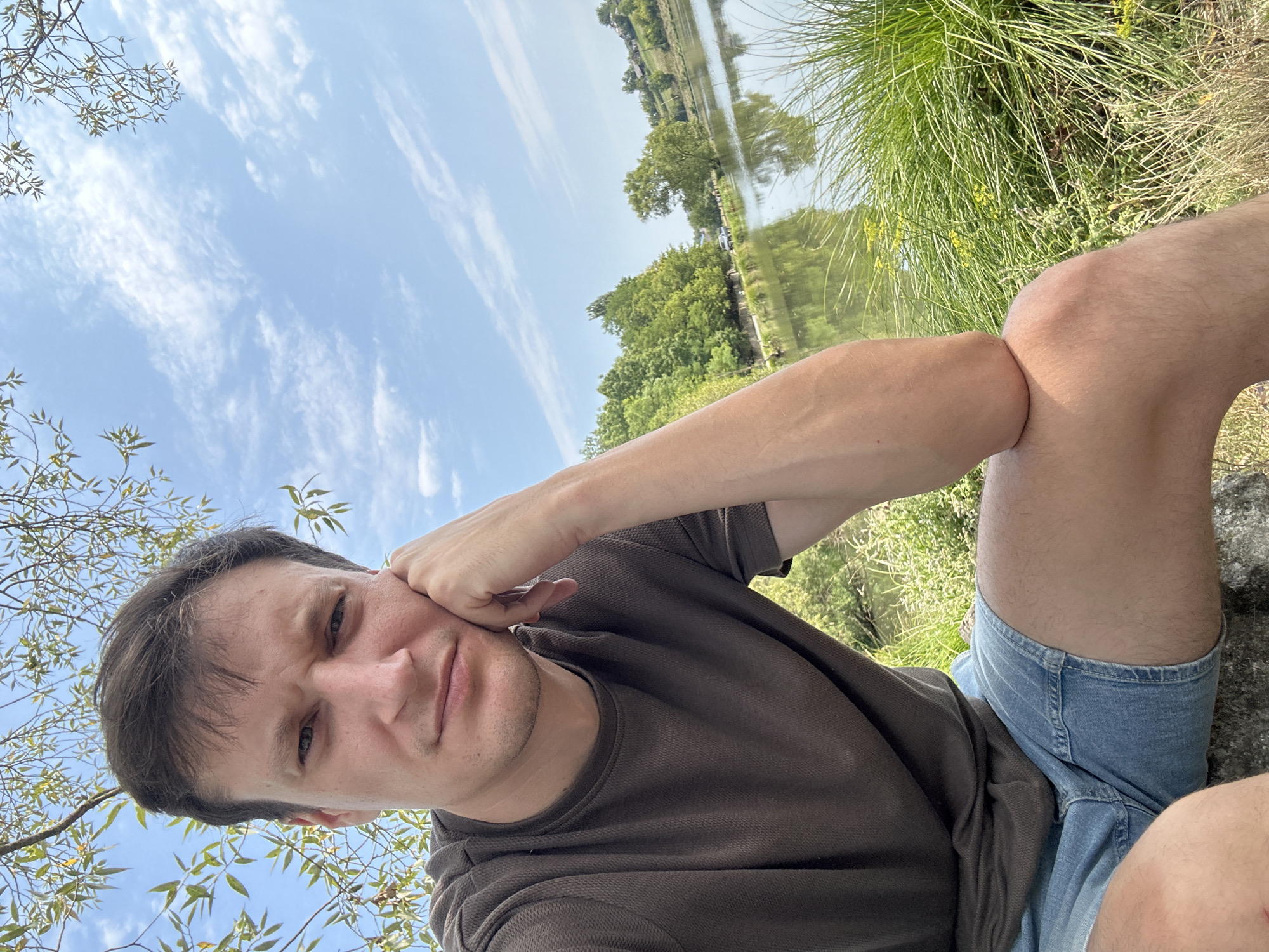 A medium shot captures a man with brown hair, wearing a dark brown t-shirt and blue denim shorts, seated outdoors. He is leaning on his right hand, with his chin resting on his knuckles, looking towards the camera with a pensive expression. The background features a body of water, lush green trees and grass, and a bright blue sky with scattered white clouds. The man's pose suggests a moment of reflection or relaxation. The scene is set in a natural environment, possibly near a lake or river. The clothing and scenery indicate a warm, sunny day. The specific location in the city of Makiv, Ukraine is not discernible from the image itself.