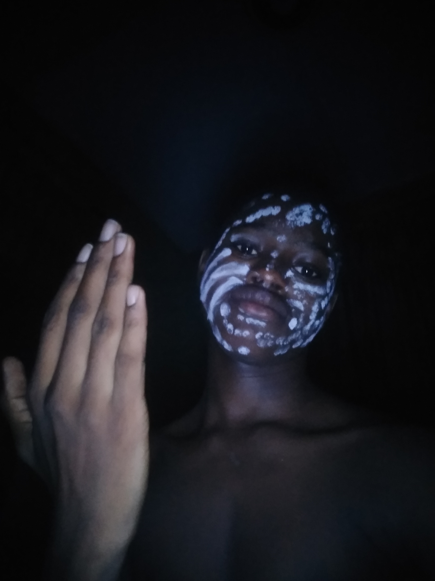 A dimly lit close-up photo shows a dark-skinned person with white paint designs on their face. The designs include stripes and dots. The person's bare chest and outstretched left hand are visible. The background is dark. The location is specified as Shangisha, Nigeria.