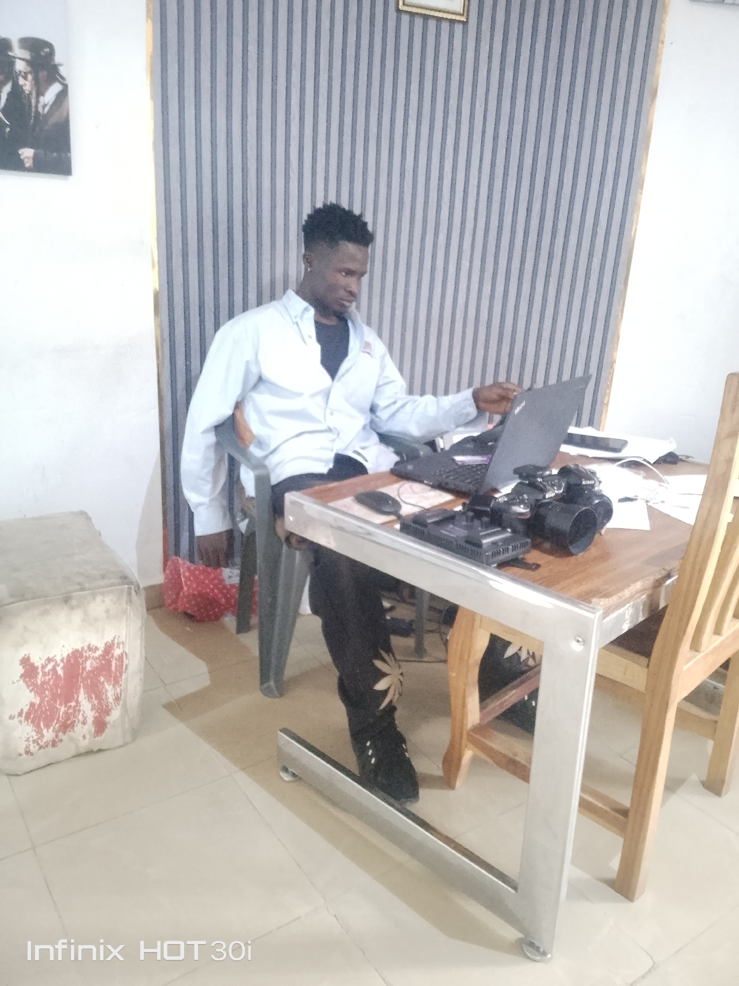A person is sitting at a desk in front of a laptop. The person is wearing a light blue collared shirt and black pants with a white floral pattern. They are looking down at the laptop, which is open. On the desk, there is a black camera, a computer mouse, and some papers. A wooden chair is next to the desk. In the background, there is a wall with vertical stripes and a framed picture on the wall. To the left of the desk, there is a cube-shaped stool with red and white markings. The setting appears to be indoors, possibly an office or a room in a home. The lighting suggests it might be daytime. The text "Infinix HOT30i" is visible at the bottom left of the image.
