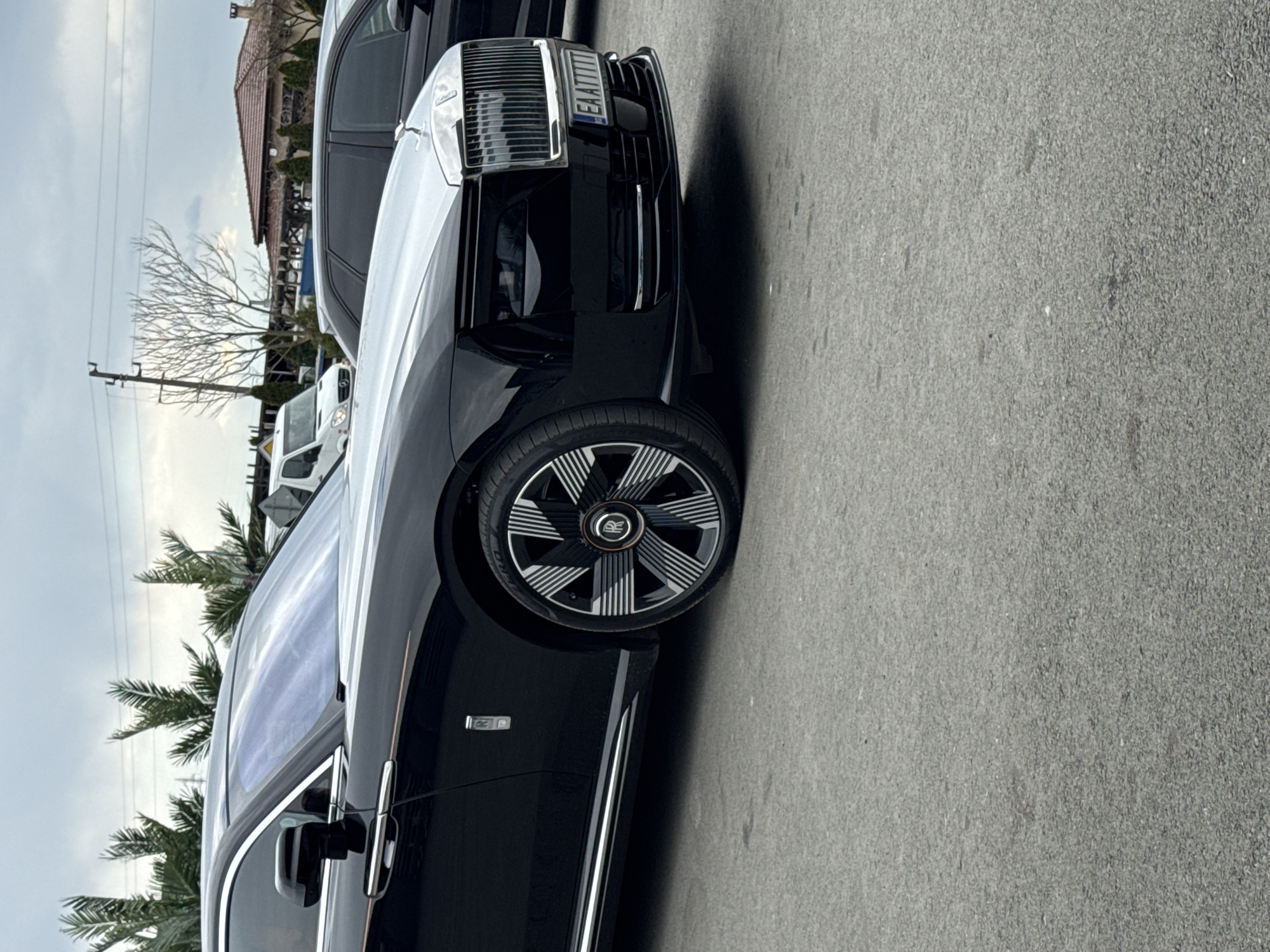 The image shows the front of a black Rolls-Royce car parked on a paved surface, likely a road or parking lot. The front grille and wheel with the Rolls-Royce logo are visible. The license plate reads "EA 7177 TT". Palm trees and a building are visible in the background. The location is inferred to be in Yambol, Bulgaria.