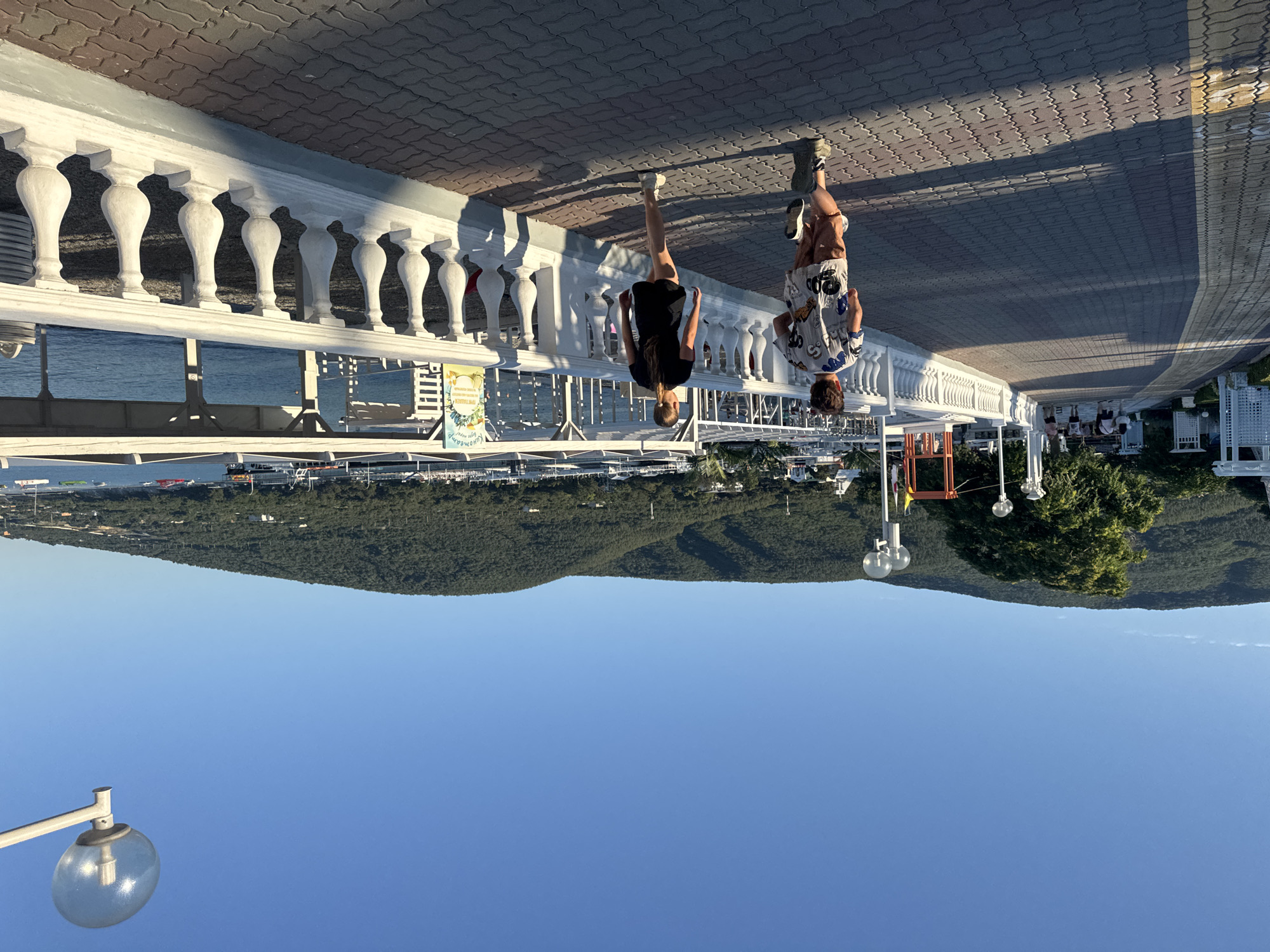 Two children, a boy and a girl, are running on a paved promenade along the waterfront in an unknown city. The boy, in the foreground, is wearing a patterned t-shirt and shorts, while the girl is wearing a black t-shirt and shorts. They are running away from the camera. To their right is a white balustrade separating the promenade from the water, where several boats are docked. In the background, there are green, tree-covered hills under a clear blue sky. The promenade is made of red and gray interlocking pavers, and is lined with white decorative railings and lampposts. Sunlight casts long shadows on the promenade. There are other people walking in the distance on the left side of the promenade, and white structures, possibly buildings or shelters, are visible along the waterfront.