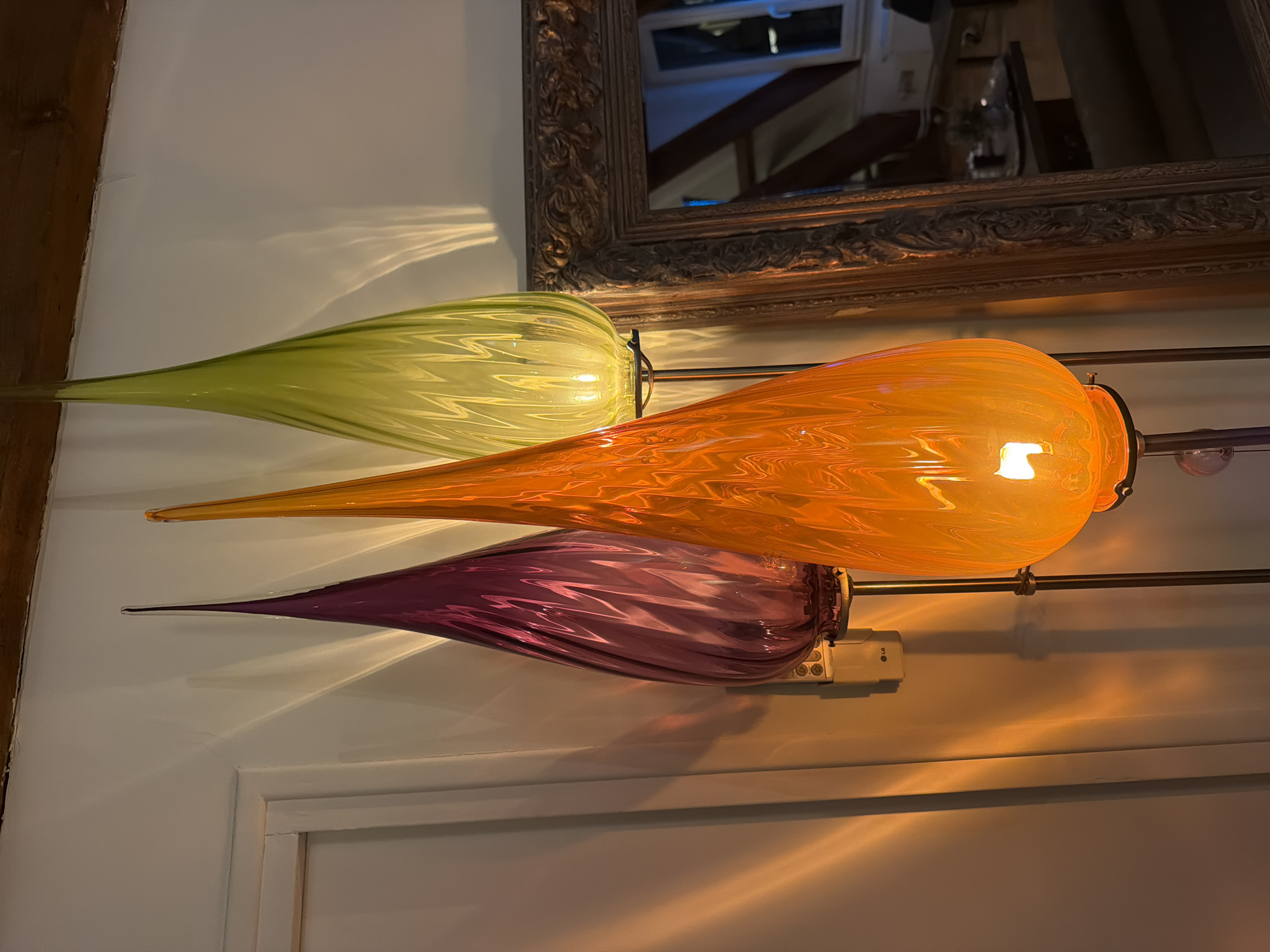 This indoor scene is located within a residence in Paris, France. Three tall, teardrop-shaped glass floor lamps stand in a row. From left to right, the lamps are purple/magenta, orange, and lime green. Each lamp is ribbed or swirled in texture, illuminated from within, and mounted on a metallic pole. The lamps emit warm light, casting elongated, wavy patterns onto the adjacent white wall.
To the left of the purple lamp, a white-framed doorway is partially visible against the white wall. Above, a dark, rough-hewn wooden beam runs horizontally across the top of the frame. Between the purple and orange lamps, a small, off-white rectangular wall-mounted device, labeled "LG" and featuring several circular buttons, is affixed to the wall.
To the right, a large, ornate, gold-framed mirror leans against the wall, reflecting a portion of the room beyond the lamps. The reflection in the mirror shows a window with a white frame, another dark wooden beam in the ceiling, and what appears to be a table with several objects, including a dark tray, a small plant, and other indistinct items. The scene is illuminated by the artificial light from the lamps. No people are present.