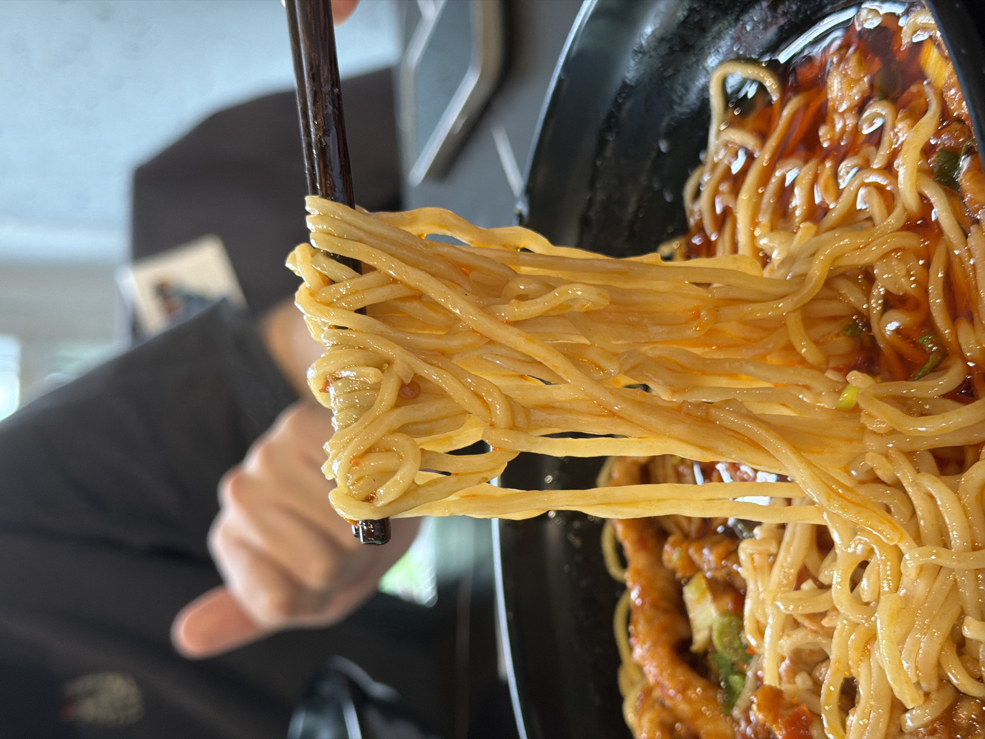 A close-up shot depicts a person using chopsticks to lift a portion of noodles from a black bowl, presumably containing the remainder of the dish. The noodles appear to be coated in a sauce. The background features a blurred figure of the person holding the chopsticks, dressed in a dark shirt, and a glimpse of the surrounding environment. It is not possible to determine the location of the image as Saint Petersburg, Russia.