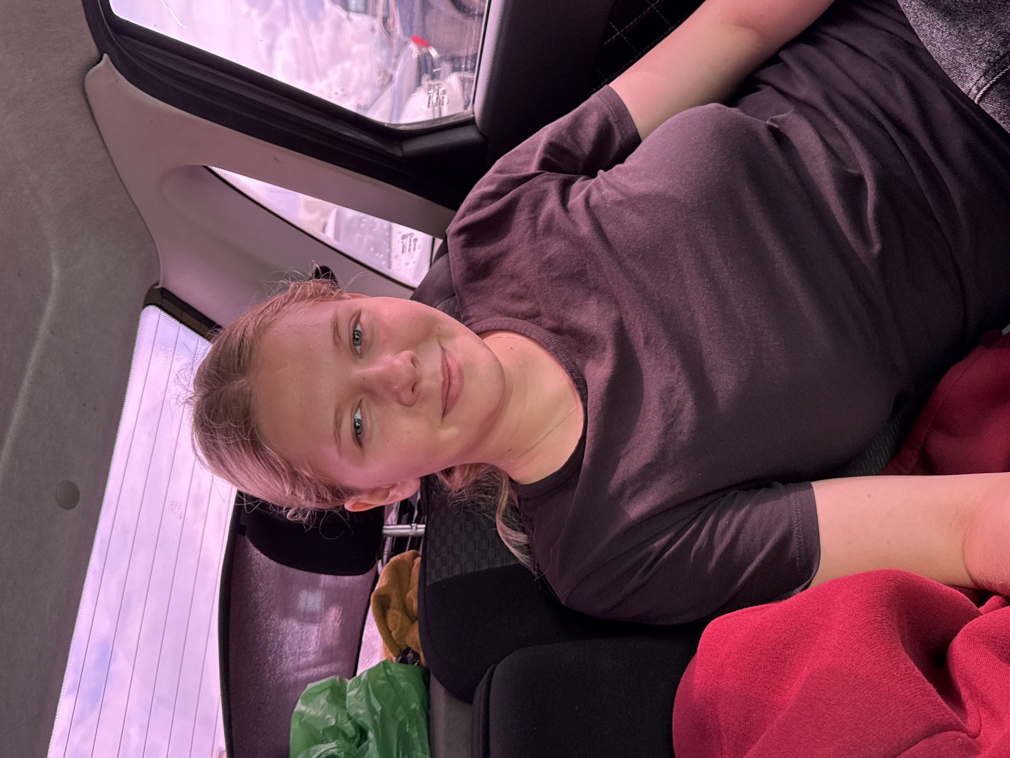 A young Caucasian female with blonde hair pulled back is seated in the back seat of a vehicle, looking directly at the camera. She is wearing a dark grey t-shirt. To her left, a bright red fabric is visible, and behind her, a green plastic bag and a brown item are partially visible. The vehicle's interior, including the back seat and ceiling, is discernible. Through the car windows, a pinkish sky and the blurry outlines of other vehicles are visible. The background suggests an outdoor setting, possibly a parking lot or road.
