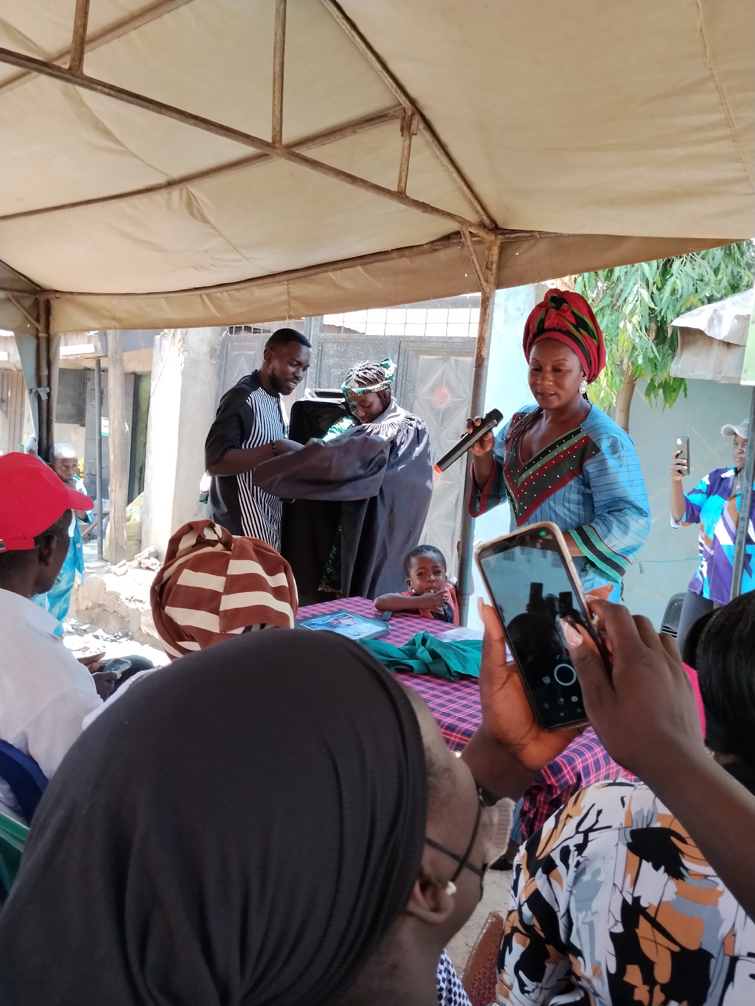 A group of people are gathered outdoors under a white canopy, which is supported by metal poles. The location is Sabon Tasha, Nigeria. A woman is holding a microphone and speaking. She is wearing a blue patterned dress, a red and green head wrap, and earrings. She is standing near a table covered with a red and purple checkered tablecloth. A child is visible near the table, looking directly at the camera. A man in a striped shirt and a woman in a head covering are holding a dark-colored garment, possibly presenting it to someone. People are seated in the foreground, and some are holding up mobile phones to record the event. There are various buildings and structures visible in the background.