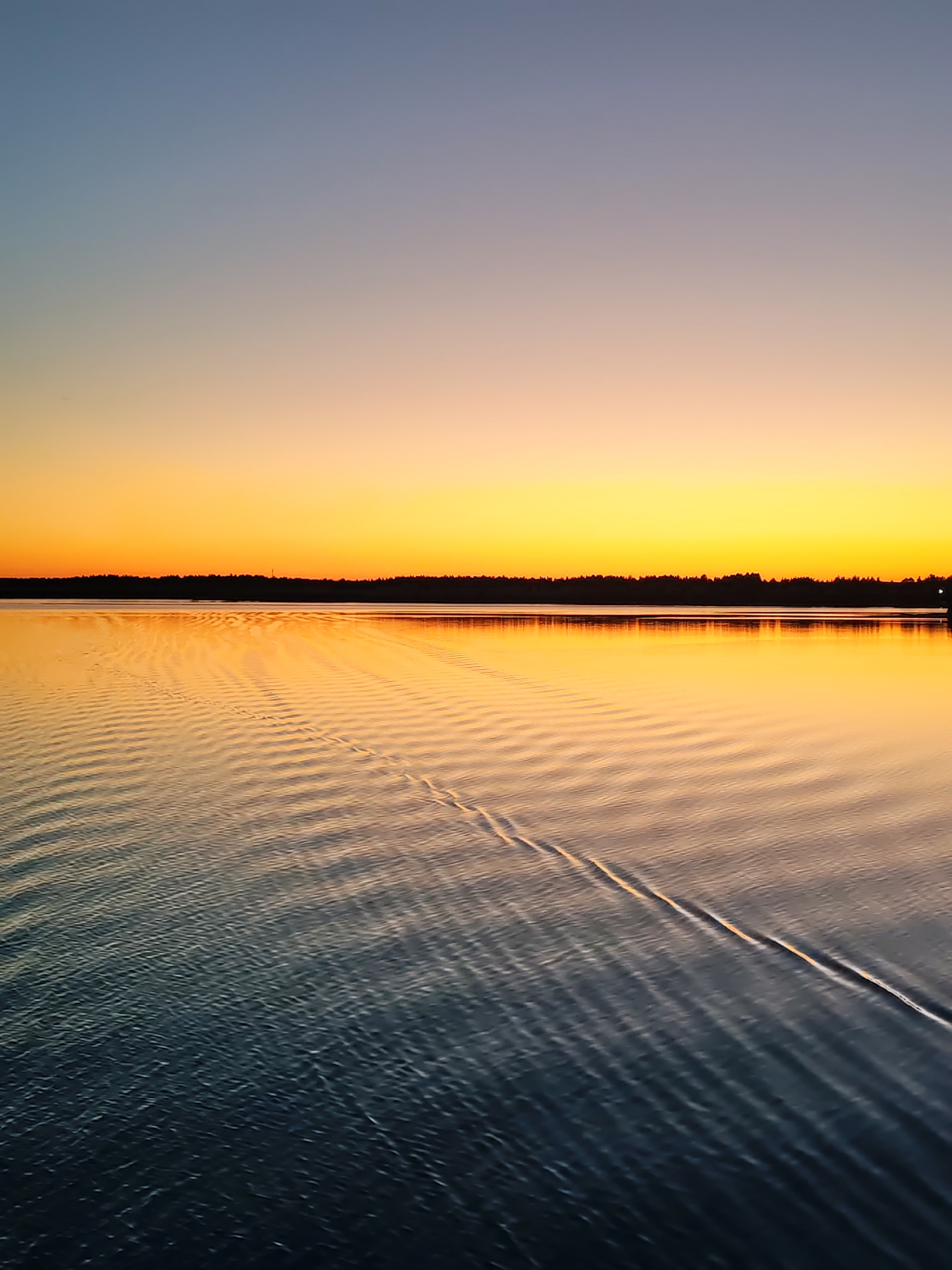 The image captures a serene landscape at either sunrise or sunset over a large body of water. The sky exhibits a distinct gradient, transitioning from a pale blue at the top to a muted gray-lavender, then broadening into vibrant bands of yellow and intense orange as it approaches the horizon. A continuous, dark silhouette of a forested landmass marks the distant horizon line, with a faint, slender vertical structure visible slightly left of center. The water's surface acts as a mirror, reflecting the warm orange and yellow light from the sky, while the foreground water deepens to darker shades of blue and gray. The entire water surface is characterized by numerous uniform, parallel ripples. A prominent, linear wake or disturbance, consisting of two distinct parallel raised lines, traverses the water diagonally from the mid-right foreground towards the upper-left midground. No human figures or discernible vessels are present. The specific location in the city of Unknown, Unknown cannot be determined from the visual information.