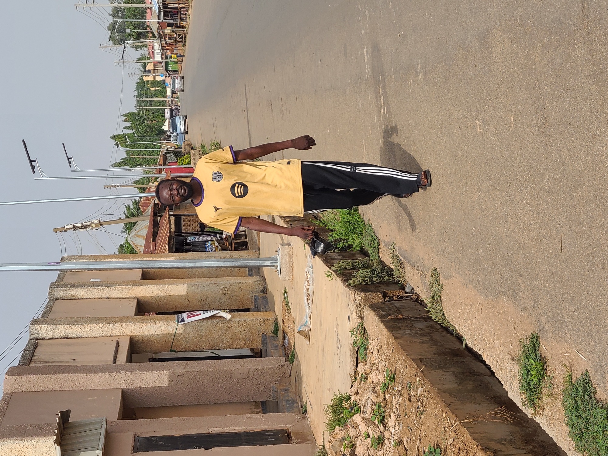 A Black man stands on the right side of a paved road in Barakin Lerawi, Nigeria, posing for a photograph. He faces slightly left towards the camera with a neutral expression, wearing a yellow short-sleeved t-shirt with purple trim, black track pants with white side stripes, and dark sandals. The t-shirt features a circular logo resembling Spotify's, and another emblem, possibly a football crest, on the upper left chest. He holds a small dark object in his left hand.
The scene is a sunlit street during the daytime, under a clear sky. The road stretches into the distance, showing light vehicle traffic. Modern solar-powered streetlights are regularly spaced along the roadside. To the left, a multi-story building with a light brown, stepped concrete facade is prominent, separated from the road by a concrete drainage ditch filled with some vegetation. Other buildings, some with corrugated roofs, are visible further down the street. Green weeds and plants grow along the ditch and road edges. Overhead power lines crisscross parts of the sky. Text, possibly "IBEFULER," is partially visible on a sign attached to the concrete building on the left.