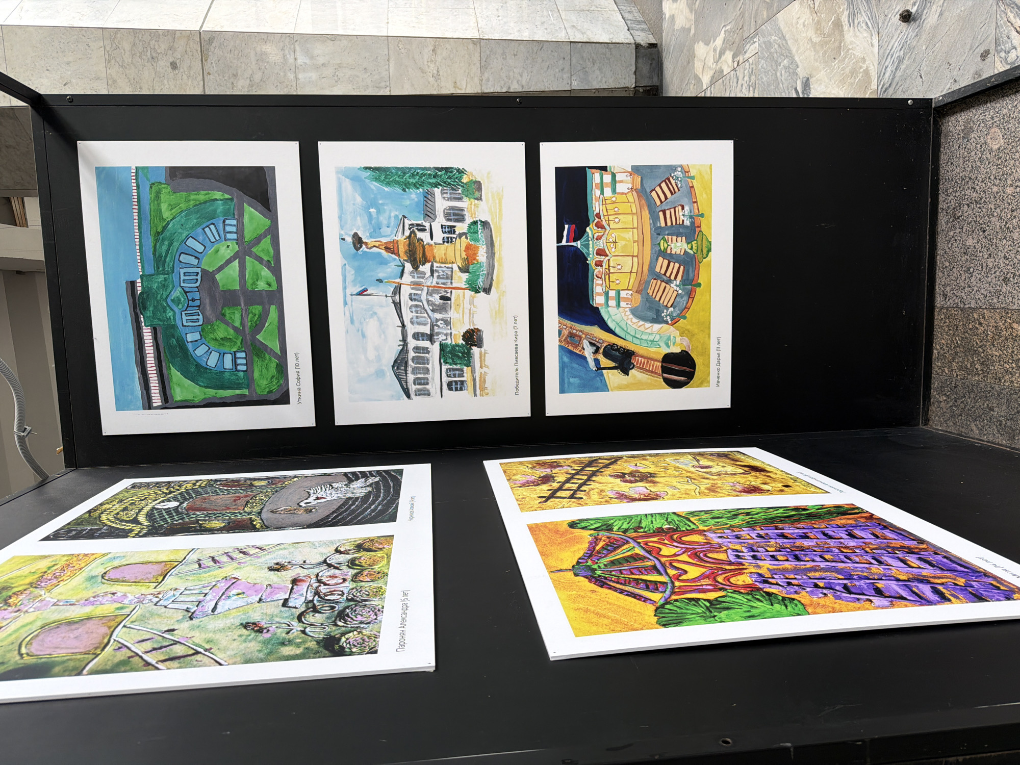 The image shows an outdoor exhibition of children's artwork. The artwork is displayed on black panels, which are part of a larger exhibition structure. The panels are mounted on a floor made of granite tiles, some of which are patterned. The background shows a building with stone facade and a grey pipe running overhead. The artwork itself consists of colorful paintings, some of which are framed and hung on the panels, while others are mounted directly. Some of the paintings depict architectural subjects and landscapes, possibly depicting scenes from Moscow, Russia, given the presence of Russian flags in some of the artworks. The lighting suggests it is daytime. There are no people visible in the image.