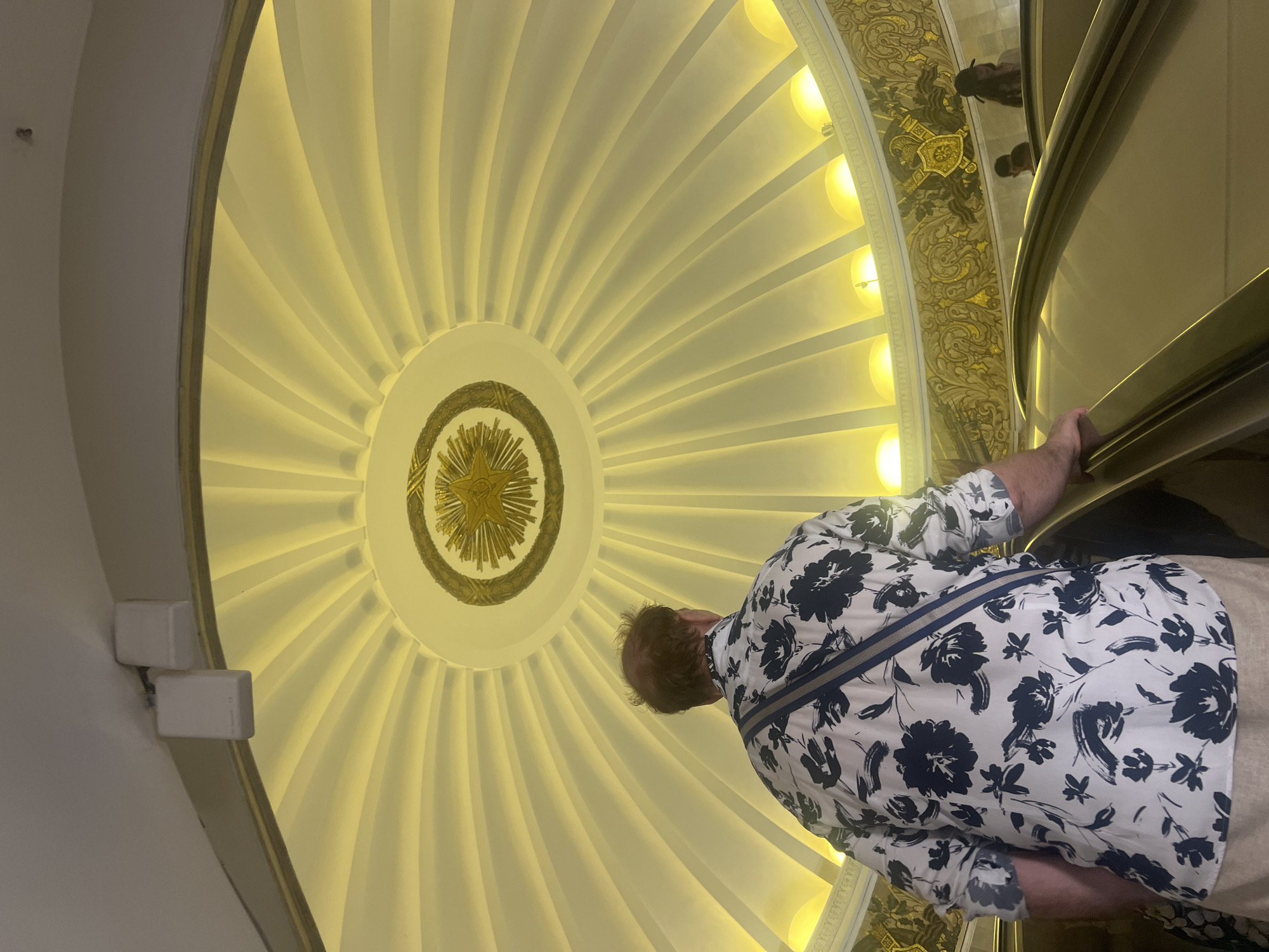 The image shows a man on an escalator looking up at a domed ceiling in what appears to be a Moscow Metro station. The ceiling is illuminated by warm yellow lights and features a circular golden emblem at its center, surrounded by radiating panels. The man is wearing a white shirt with a dark floral pattern and a blue and white strap across his chest. Other people are visible on escalators in the background, and decorative friezes with gold detailing adorn the walls. The architecture suggests the grandeur often found in Moscow's historic metro stations.