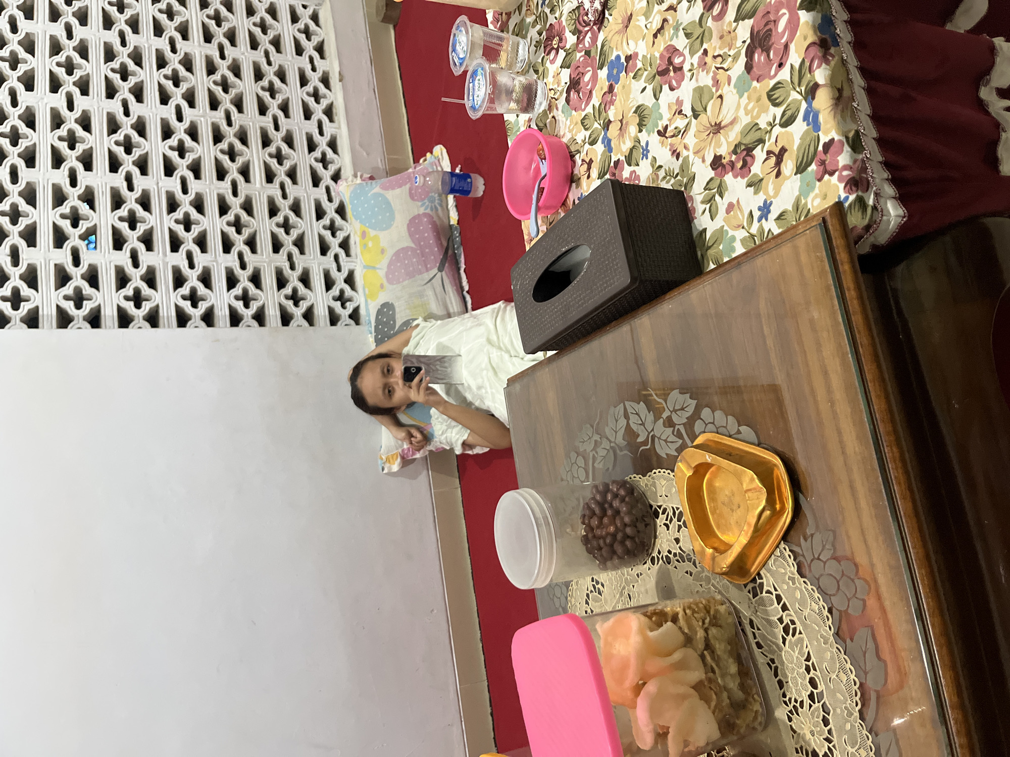 A young girl is lying on a red carpet, resting her head on a pillow with a butterfly pattern. She is holding a smartphone in her right hand, seemingly taking a selfie. She is wearing a white outfit. In the foreground, a wooden table is visible, covered with a floral tablecloth. On the table, there are several items, including a pink plastic container with what appears to be pink snacks, a transparent jar filled with small brown candies, a golden ashtray, a brown tissue box, a pink bowl with a spoon and some red berries, and two plastic cups with straws, one filled with a clear liquid. A bottle of water is also on the carpet. The background features a white wall and a decorative lattice-like structure with a cross pattern. The scene appears to be indoors, likely in a residential setting in Glagah Kulon, Indonesia, possibly a living room or a dining area.