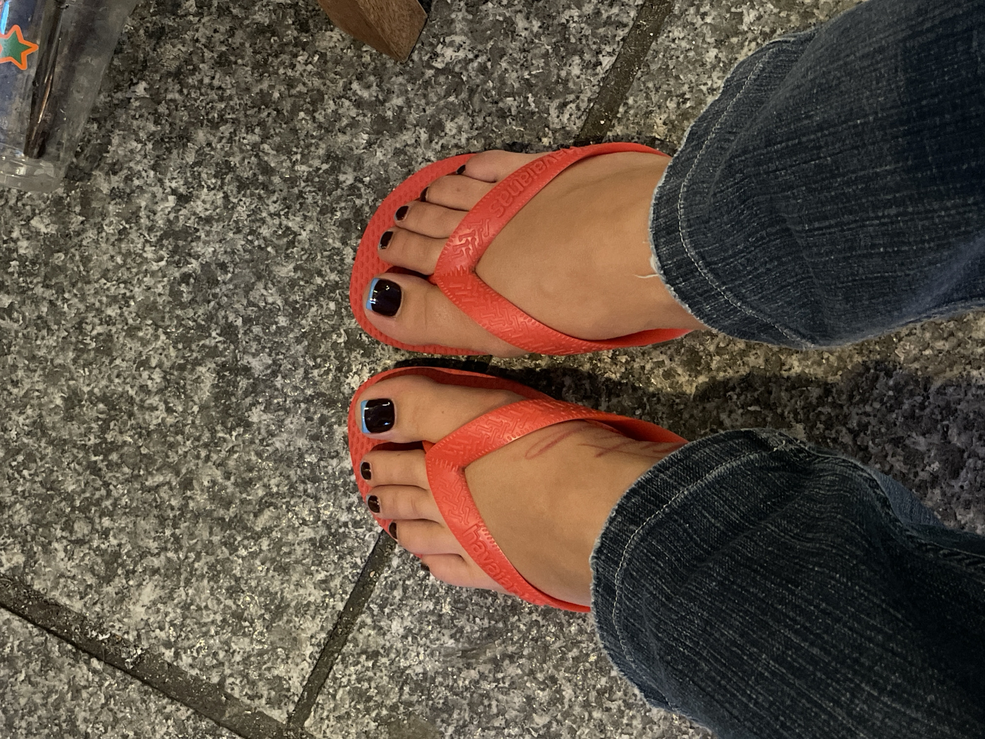 An eye-level close-up shows a pair of feet in orange Havaianas flip-flops on a gray, granite-like surface with dark grout lines. The feet are angled toward the camera. The toenails are painted a dark color. The left big toe has a thin light blue line along its edge. The person is wearing dark blue jeans. A blurred object is visible in the upper left corner. The location is implied to be Paris, France.