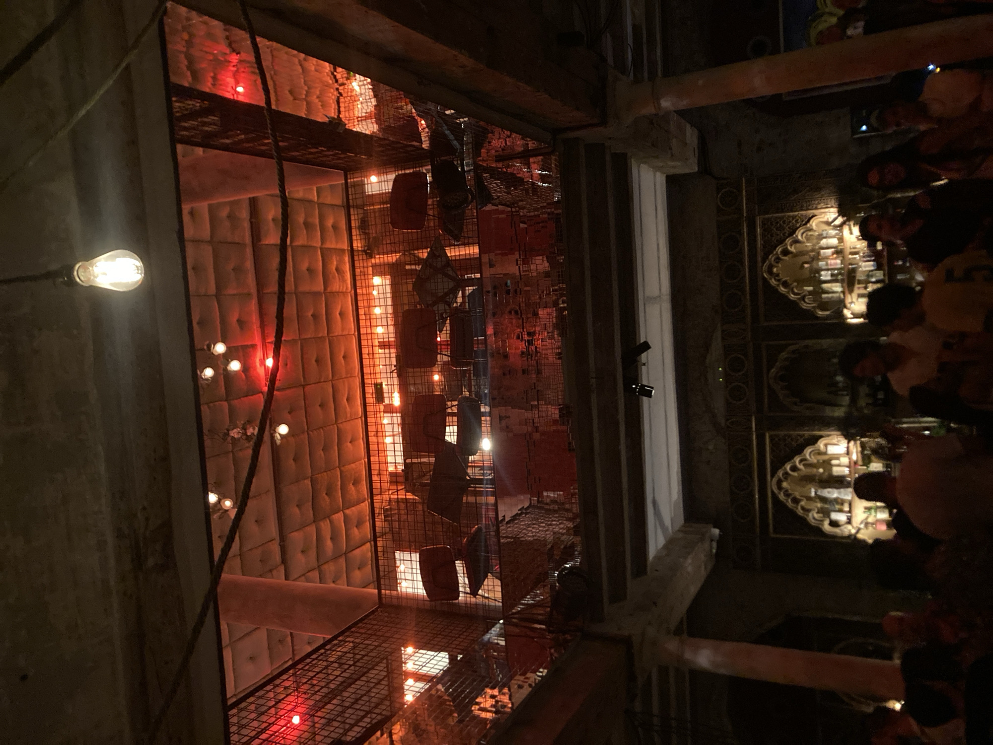 The image depicts the interior of a dimly lit establishment with a multi-level design, likely a bar or restaurant, located in Barcelona, Spain. Red and warm lighting illuminates the scene, highlighting architectural features and creating an intimate atmosphere.
On the upper level, a metal-grated balcony overlooks the lower area. Several chairs are visible on this level, arranged around tables that are partially obscured by the grating. The ceiling above this level features a quilted, padded texture, illuminated by strategically placed lights and what appear to be string lights. A prominent Edison bulb hangs from a cord in the foreground.
The lower level reveals a bar area with a decorative, arched facade. Bottles of liquor are displayed on shelves behind the bar. Several individuals are gathered in this area, some facing the bar and others interacting with each other. They appear to be patrons of the establishment. The architecture includes stone pillars and decorative elements, suggesting a possibly historic or elaborately designed space. The overall impression is of a lively, social environment with a distinct, moody ambiance.