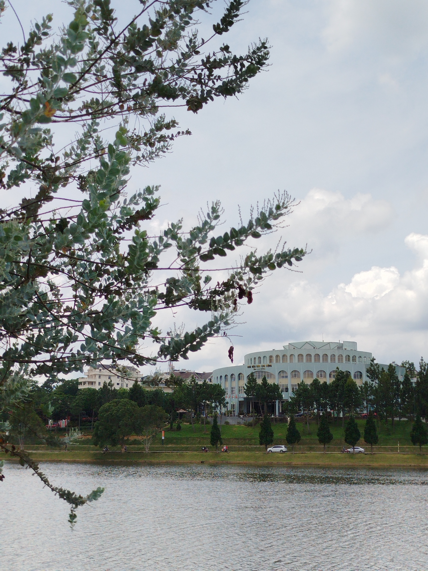 The image shows a body of water in the foreground, with a tree branch in the upper left corner obscuring part of the view. In the middle ground, there is a grassy embankment with a road and several people on motorcycles. Beyond the embankment, there are trees and buildings, including a prominent light blue, multi-story building with a curved facade and many arched windows. The sky is cloudy. The building in the background is identified as the Da Lat Workers' Cultural Palace (Nhà Văn Hóa Lao Động Tỉnh Lâm Đồng), a significant landmark in Da Lat, Vietnam, situated near Xuan Huong Lake.