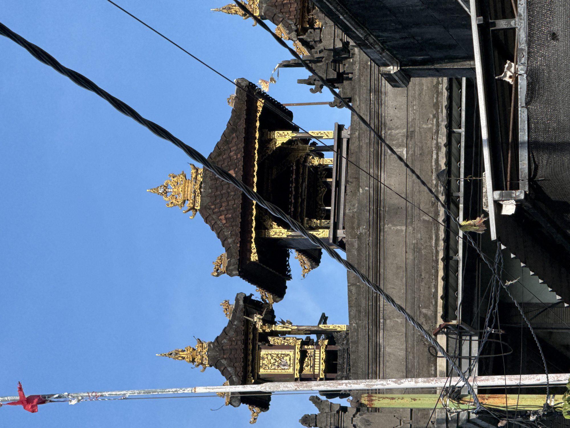 The image captures a vibrant overhead view of traditional Balinese architecture in Kuta, Indonesia, set against a clear, bright blue sky. The scene is illuminated by daylight, suggesting mid-morning or afternoon.
The primary subjects are two ornate structures, likely parts of a temple or traditional building. They feature distinctive multi-tiered roofs made of dark, reddish-brown tiles. These roofs and the structures beneath them are richly adorned with intricate golden carvings and decorative elements, including pointed spires and detailed patterns along the edges and facades. The golden ornamentation stands out brilliantly against the dark building materials and the sky.
A prominent feature of the foreground and mid-ground is a network of electrical or communication cables. A thick, twisted black cable runs diagonally across the upper left part of the image, while several thinner, tangled wires are visible on the right, intersecting with a more utilitarian concrete building. This building has visible metal frames, scaffolding, and black mesh netting, contrasting with the traditional aesthetic of the main structures.
On the far left, a white pole with a small, bright red fabric tied to it can be seen, possibly a decoration or small flag. Other small, light-colored fabric pieces are also visible, fluttering near the roofs and cables.
No human activity is depicted; the scene focuses on the architectural and urban landscape. There is no visible text. The overall impression is a blend of rich cultural heritage with the realities of modern infrastructure in a tropical, sunny setting.