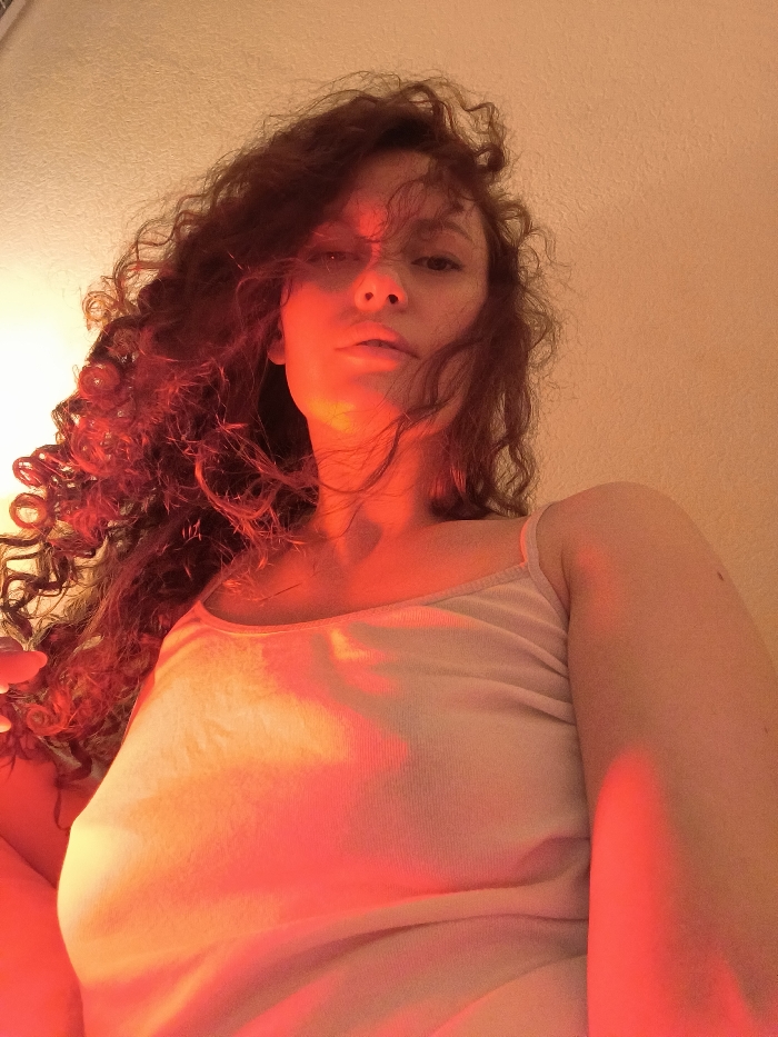 An individual, identified as female, with voluminous, dark, curly hair, is depicted from a low-angle perspective in an indoor environment. She is wearing a light-colored, possibly off-white, spaghetti-strap top. Her head is angled slightly downward, and her gaze is directed towards the camera with a neutral facial expression. The scene is illuminated by strong artificial lighting, predominantly red and orange in hue, creating a dramatic ambiance. A concentrated red light source appears to originate from below and to the viewer's left, intensely illuminating the left side of her face, neck, and hair, as well as parts of her upper torso. The background is a plain, light-colored wall or ceiling surface, lacking any discernible features. Visual elements in the image do not provide specific identifiers to determine a location within Saint Petersburg, Russia.