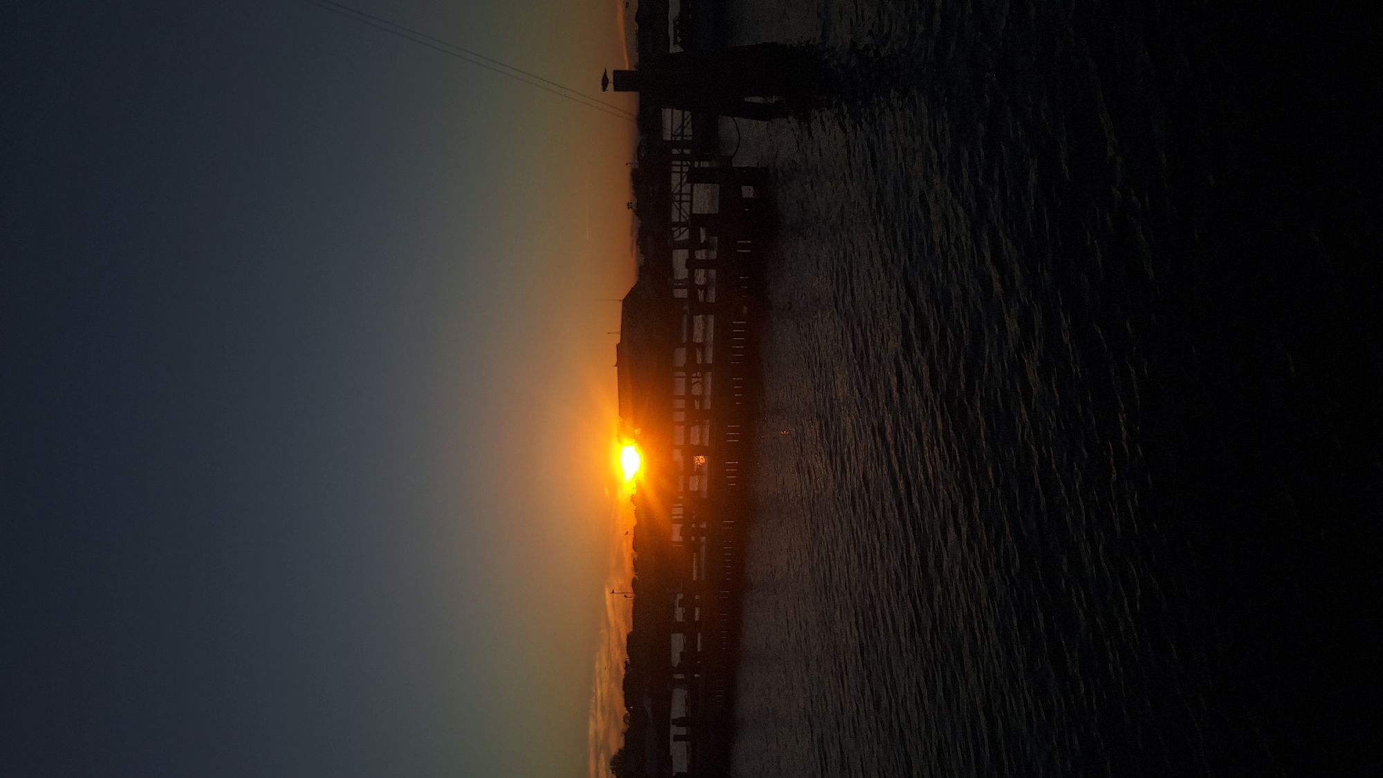 A sunset is captured over the water in Nantucket, Massachusetts, United States of America. The sun is low on the horizon, casting a warm glow across the sky, and its rays are visible behind a pier. The water is dark and choppy, with subtle reflections of the sunset. The pier extends into the water, with silhouetted structures and posts visible. A bird is perched on a post to the right of the pier. The sky above is a gradient of dark blue to a lighter orange and yellow near the horizon.