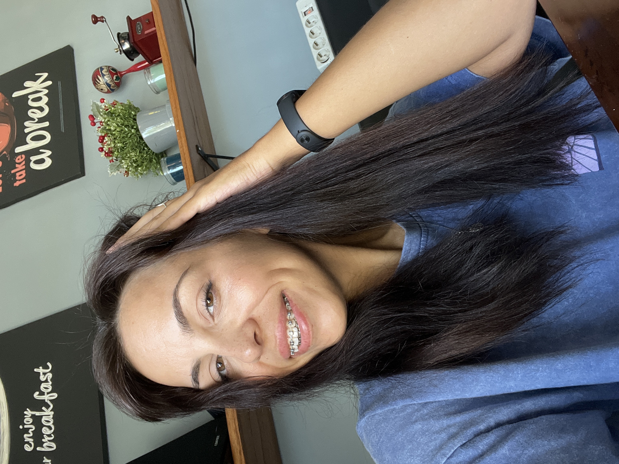 A woman with braces smiles at the camera, holding her long, dark brown hair. She wears a blue t-shirt and a black fitness tracker on her wrist. In the background, there is a shelf with decorative items, including a sign that says "take a break" and another that says "enjoy your breakfast." A power outlet strip is visible on the wall behind her arm. It's unlikely to ascertain the precise location in Kuta Selatan, Indonesia from the image provided.