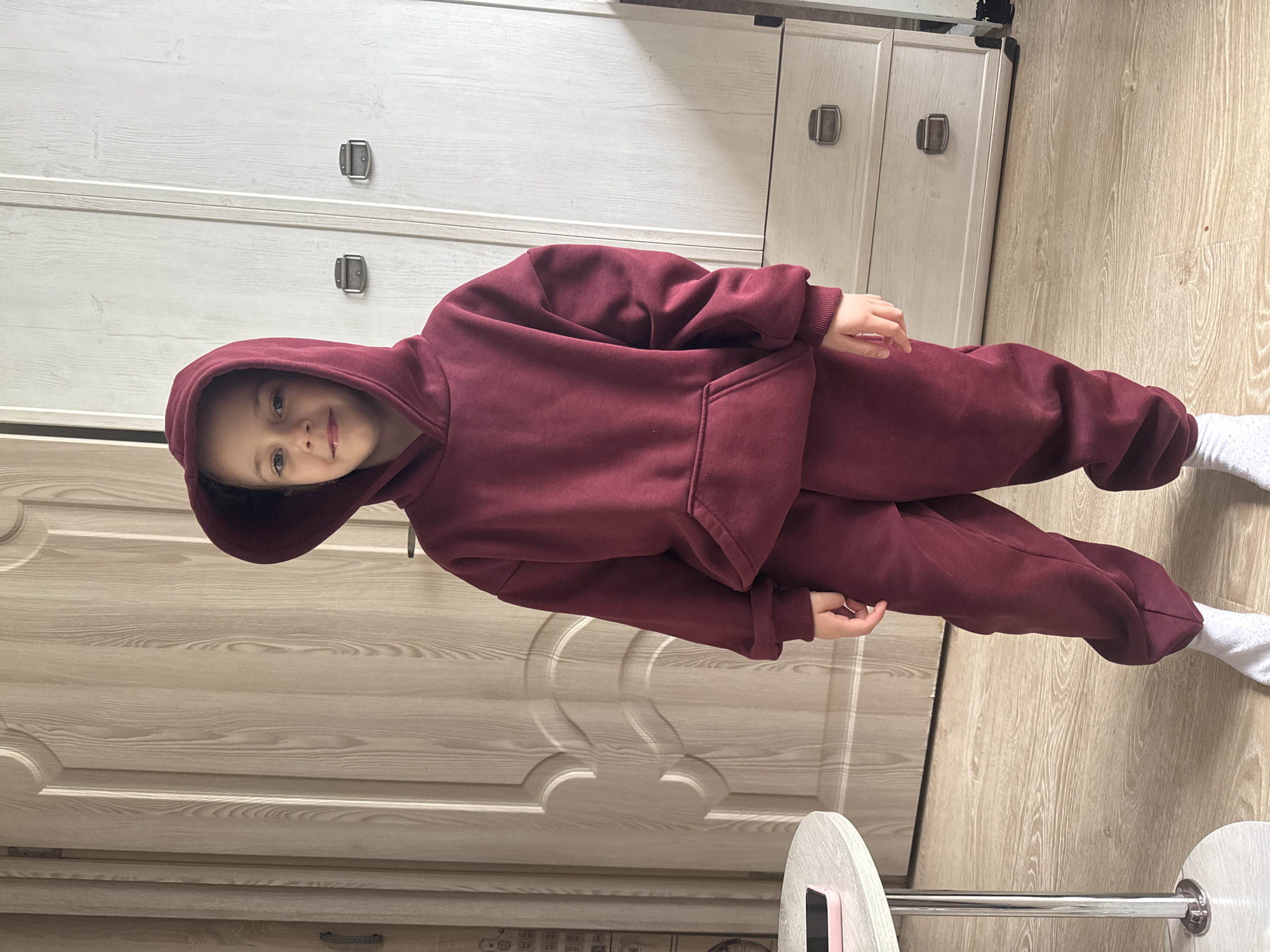 A child wearing a burgundy hoodie and matching pants stands indoors against a background of light-colored cabinets and wood-patterned flooring. The child is wearing white socks and is smiling. The location is Раздолье, Russia.