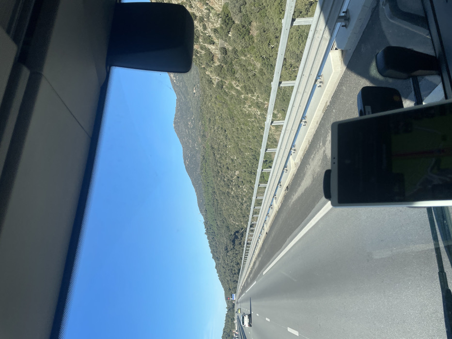 The image displays a forward-facing view from inside a vehicle traveling on a multi-lane highway in Le Perthus, France. A section of the vehicle's dark gray dashboard is visible at the bottom. Mounted centrally on the dashboard is a dark smartphone or tablet, its screen displaying a navigation application with green and red route lines. Above the device, the vehicle's clear windshield offers an unobstructed view of the highway and surrounding landscape. A dark rearview mirror housing is partially visible in the upper right corner, attached to the vehicle's ceiling lining.
The highway, constructed of asphalt, extends directly forward and slightly to the left, marked with clear white solid and dashed lane lines. Several distant vehicles, including a distinct white car, are visible in the left lane, heading away from the viewer. Along the right side of the highway, a metal guardrail runs parallel to the direction of travel, supported by concrete barriers. Beyond this guardrail, a steep mountainside covered in dense green vegetation descends, while a higher, rockier mountain peak is visible further in the background. The sky above is clear and bright blue, indicating sunny weather conditions.
