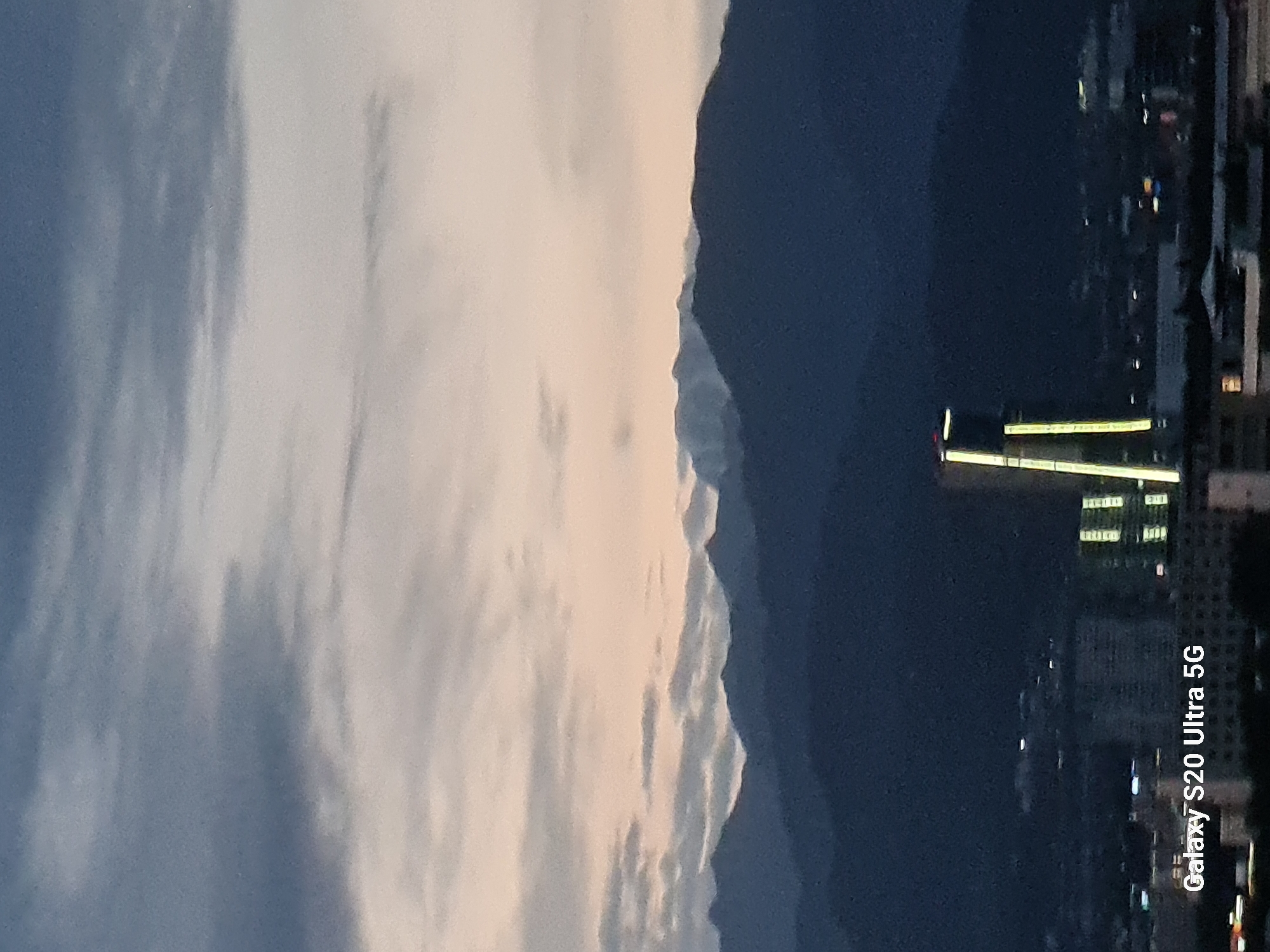This is a vertical photograph of a city skyline at dusk, with a mountainous backdrop. The left side of the image features a sky filled with soft, diffused clouds that are beginning to catch the warm, golden hues of the setting sun. This celestial display transitions into a dark, silhouetted mountain range that forms the central feature of the image. To the right of the mountains, the city's lights begin to twinkle, indicating a transition from day to night. The cityscape is partially obscured by the darkness, but illuminated structures, including a prominent building with bright, horizontal lights, are visible. The overall atmosphere is serene and transitional, capturing the quiet beauty of day yielding to night. The time of day is dusk or twilight, with the sky transitioning from a lighter, warmer tone to a deep, dark blue. The presence of the city skyline and the distinct mountain range suggest an urban environment nestled within a natural landscape. There is no clear indication of specific weather conditions beyond the cloudy sky. In the bottom right corner, the text "Galaxy S20 Ultra 5G" is visible, indicating the device used to capture the image.