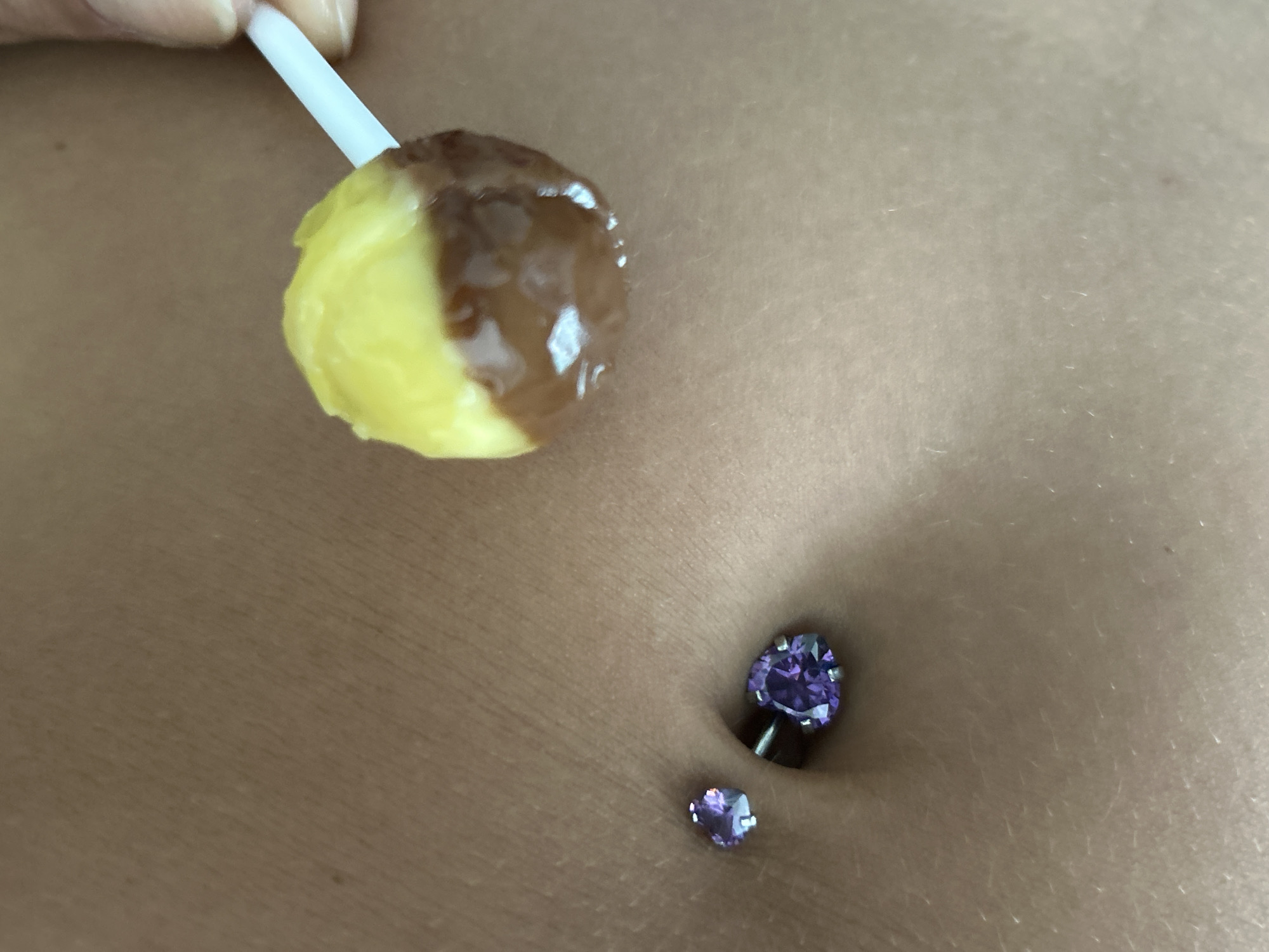 The image shows a close-up view of a person's belly button, which is adorned with two purple heart-shaped belly piercings. A lollipop, partially dipped in chocolate, is being held near the belly button. The skin appears to be light-skinned. The background is a solid, unidentifiable color, suggesting an indoor setting. There are no discernible features that would indicate a specific location in Kathu, Thailand.