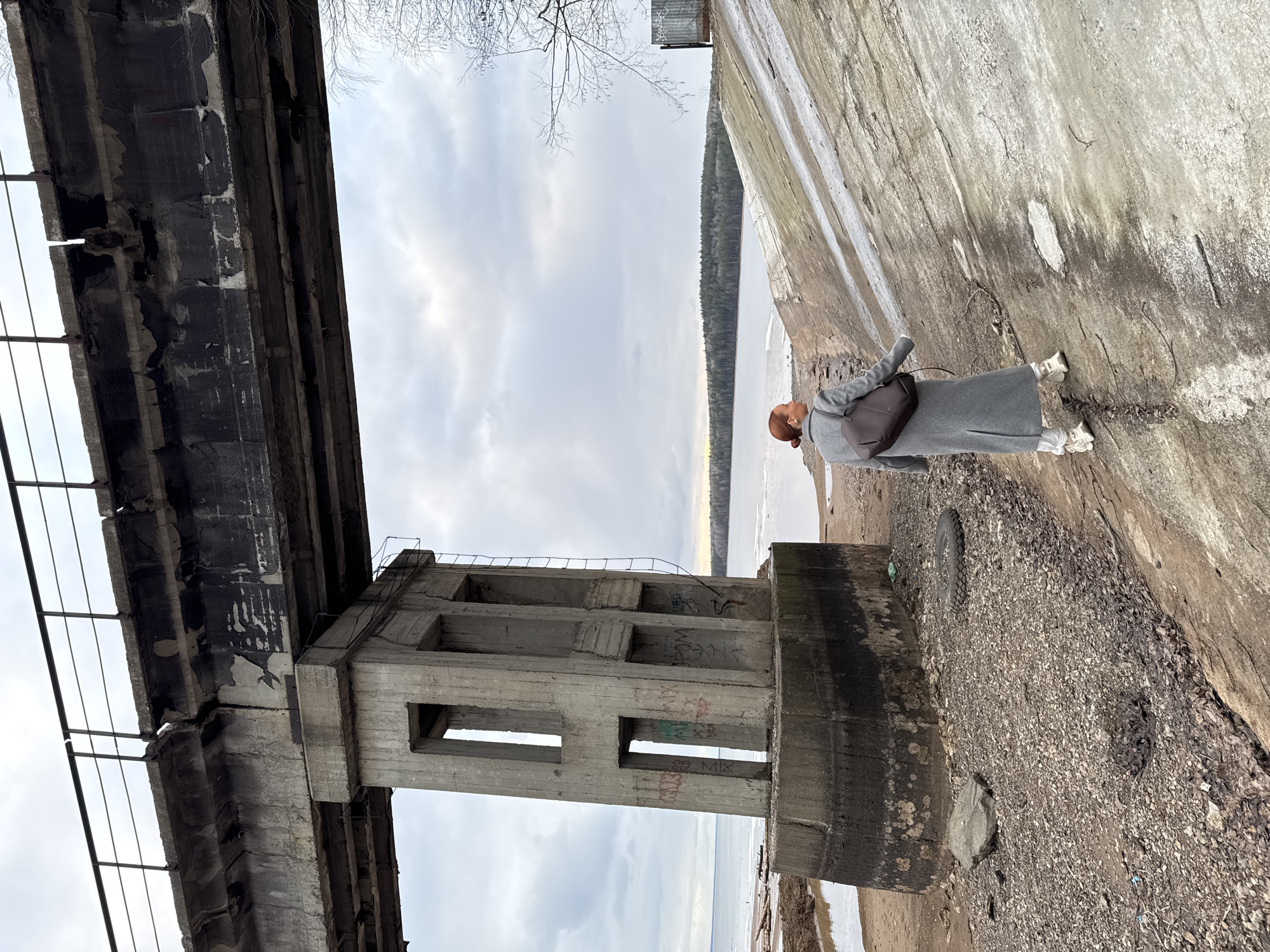 The image captures an outdoor scene in Chaikovsky, Russia, featuring a person amidst large, weathered concrete structures by a body of water under an overcast sky.
In the midground, a person stands on a rocky and gravelly bank, facing away from the viewer and looking towards the water. They are dressed in a long, dark grey coat, white sneakers, and carry a dark backpack. Their reddish-brown hair is pulled back.
To the left, a massive concrete bridge or pier structure extends, showing signs of age with dark, peeling surfaces and visible metal railings. To the right, a large, tall concrete pillar or support structure stands prominently. This pillar features several rectangular openings and is covered with graffiti, including the discernible word "MIX" and other less distinct writings in red and black. The concrete surfaces throughout the scene exhibit wear, cracks, and discoloration.
Beyond the immediate foreground, a wide expanse of water, likely a river or lake, stretches into the distance. Its surface appears calm and grey, with some brighter, possibly icy, sections closer to the bank. A distant shoreline is visible, densely forested with dark trees. The sky is entirely covered with grey clouds, indicating an overcast day without direct sunlight. Bare tree branches are visible against the sky on the upper left. On the distant concrete wall, a small, squared metal structure can be seen.
The scene suggests a cool or cold time of year, given the bare trees and muted light. The combination of imposing, decaying concrete architecture and the natural landscape of water and forest creates a stark and somewhat melancholic atmosphere.