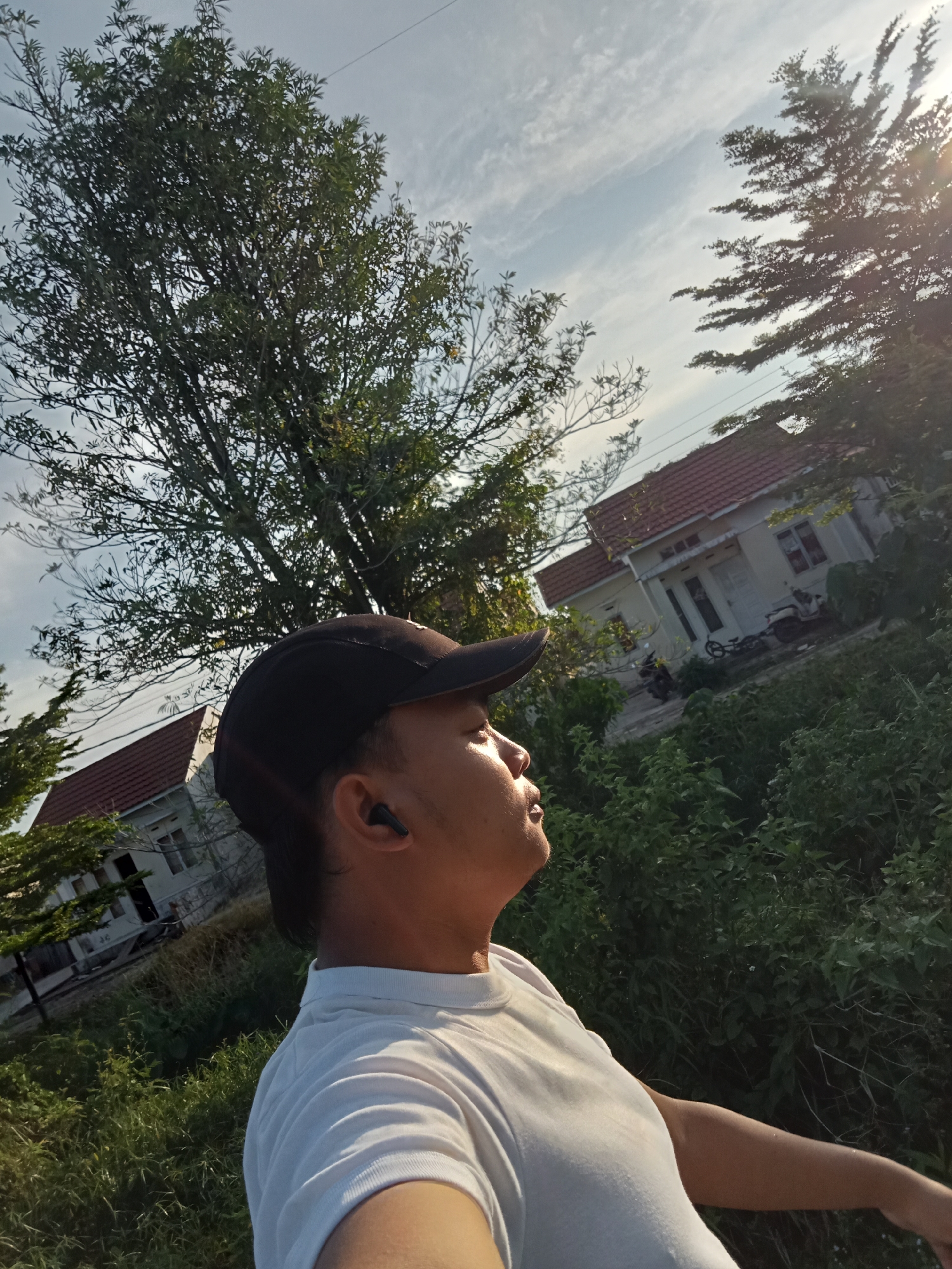 A low-angle, outdoor, eye-level, medium shot captures a person wearing a black baseball cap and a white t-shirt, looking up and to the right, outdoors in Kualu, Indonesia. The person is wearing black wireless earbuds. The background features lush green vegetation, a white house with a red tiled roof, and two motorcycles parked in front of the house. Tall trees are visible on the left and right sides of the frame, with the sun shining through the branches on the right. The sky is partly cloudy with a clear blue expanse. The image appears to have been taken during the day, possibly late afternoon, given the lighting conditions.
