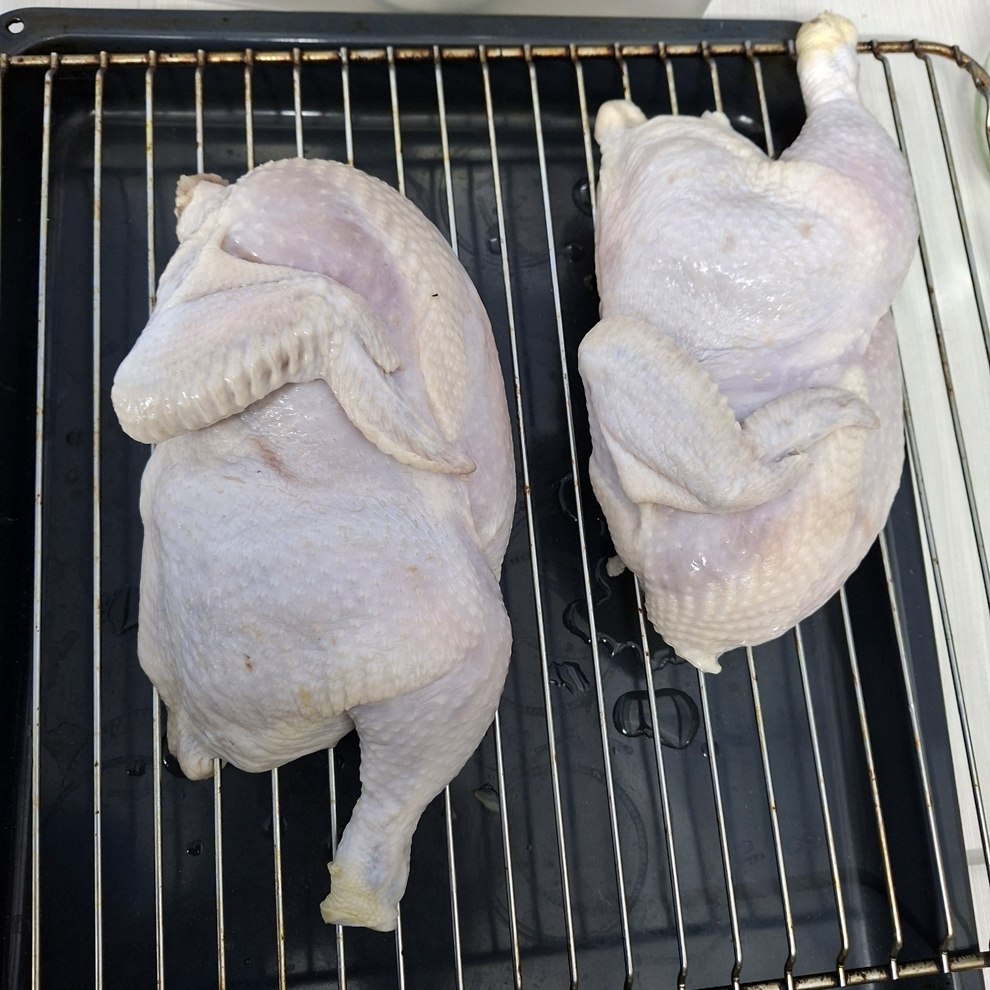 Two whole raw chickens are positioned on a metal oven rack. The chickens are pale white with visible skin texture and some pinkish undertones. They are arranged side-by-side, with the chicken on the left slightly overlapping the chicken on the right. The oven rack is placed over a dark, possibly black, baking tray which has some clear liquid droplets on its surface. The background is blurred but appears to be a light-colored surface. There are no people or any other discernible objects or details in the image. The image captures a still preparation for cooking. The location is Anapskaya, Russia.