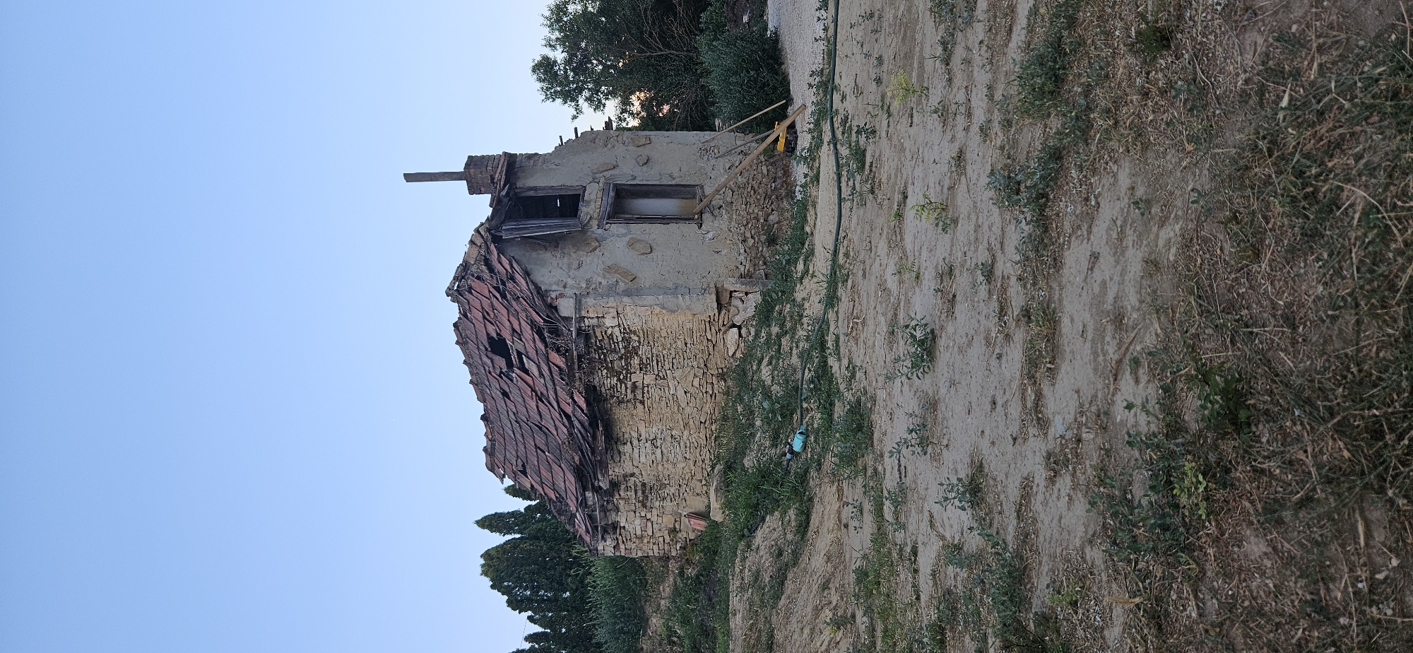 A weathered, stone and stucco structure, appearing to be an abandoned house or shed, is situated in a semi-rural or overgrown area. The building exhibits significant disrepair, with a visibly damaged roof of red tiles, exposing the rafters and interior to the elements. One section of the roof has collapsed entirely. The walls are constructed from rough-hewn stones at the base, transitioning to a lighter-colored stucco on the upper sections. There are visible openings where windows and a door would have been, some boarded up or with remnants of wooden frames. A brick chimney stands on one side of the building.
The immediate surroundings consist of uneven, sandy ground interspersed with patches of dry grass and sparse green vegetation. A green garden hose is visible on the ground, connected to a blue fitting, suggesting a water source. A red container or bucket is leaning against the side of the building. To the right of the house, leaning against the wall, are wooden planks and what appears to be a yellow toy.
Behind the structure, a line of tall, dark green coniferous trees forms a backdrop, with a wider array of green foliage to the right. The sky above is a clear, light blue, indicative of daytime, possibly late afternoon or early evening given the soft light.
The location is identified as Hévíz, Hungary. The scene conveys a sense of abandonment and neglect, with nature beginning to reclaim the structure and its surroundings. There are no people visible in the image, and no actions or interactions are depicted. The overall impression is one of decay and the passage of time.