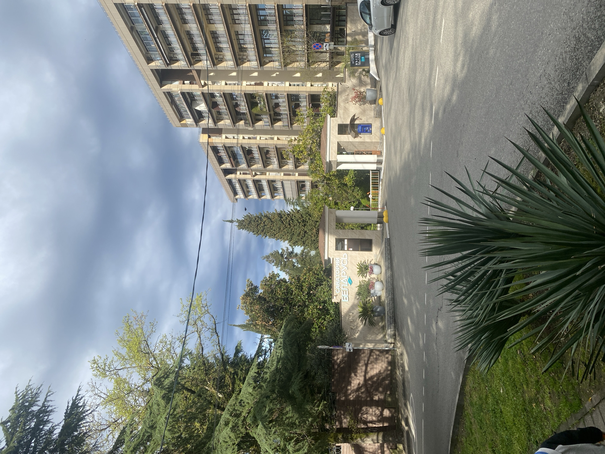 The image shows a street scene in Sochi, Russia, featuring the entrance to the "Sanatorium Belarus". A tall, multi-story building with balconies is visible on the left. The entrance to the sanatorium has a sign with its name and some plants. The street is paved with a white car parked on the side. Trees and greenery line the street. The sky is overcast.