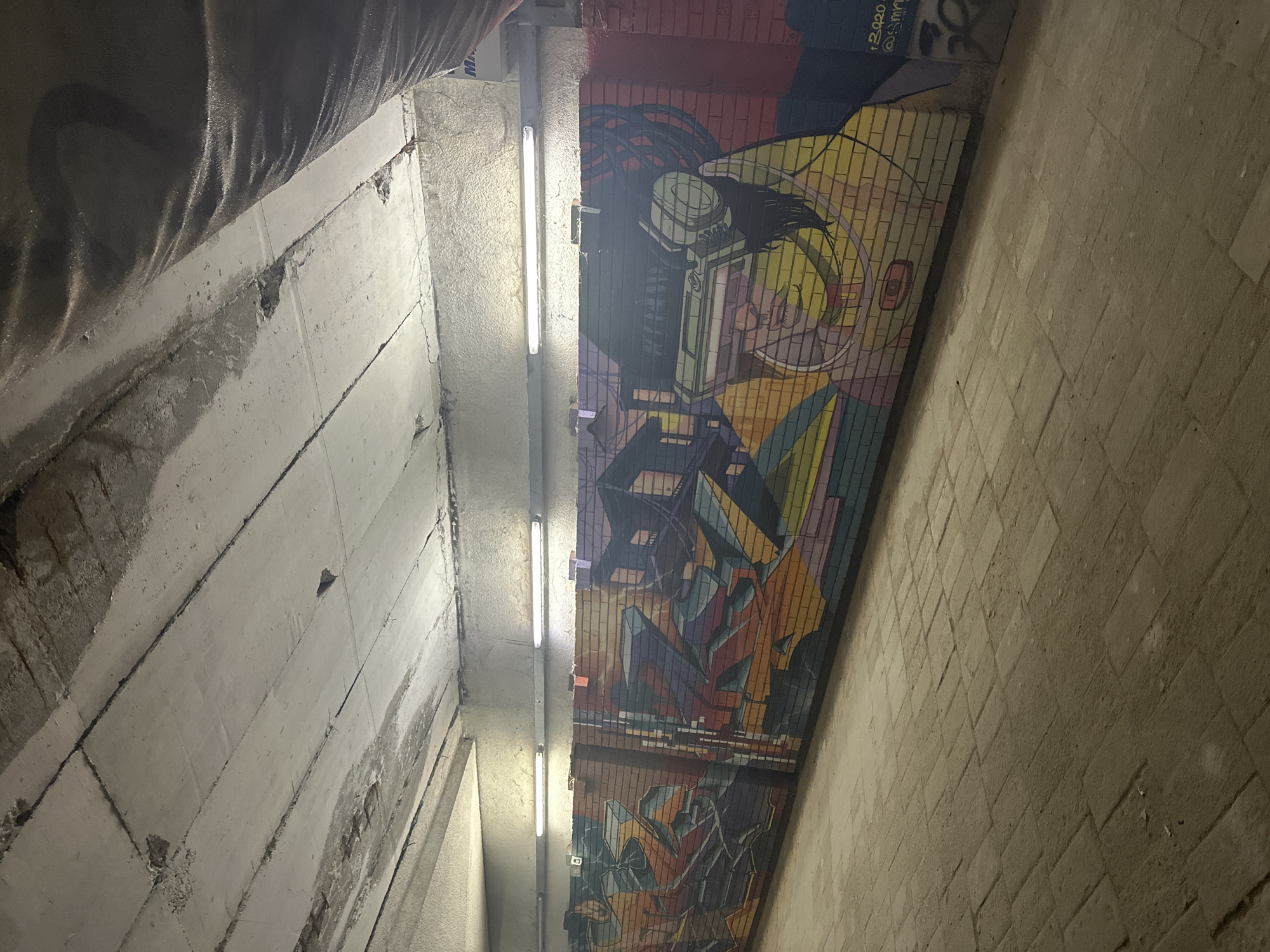 Captured from a low angle within an underpass in Sofia, Bulgaria, the image features the juxtaposition of raw concrete structures with vibrant urban art. The left side showcases the rough, segmented concrete wall, partially covered by a dark, possibly waterproof, material. Overhead, fluorescent lights cast an artificial glow onto the scene. The right side of the underpass is adorned with a colorful mural, portraying an abstract image of a face and stylized urban elements. The mural is constructed in a brick-like pattern. The underpass itself appears to be constructed with brick or stone, creating a clear demarcation between the industrial concrete and the artistic expression.